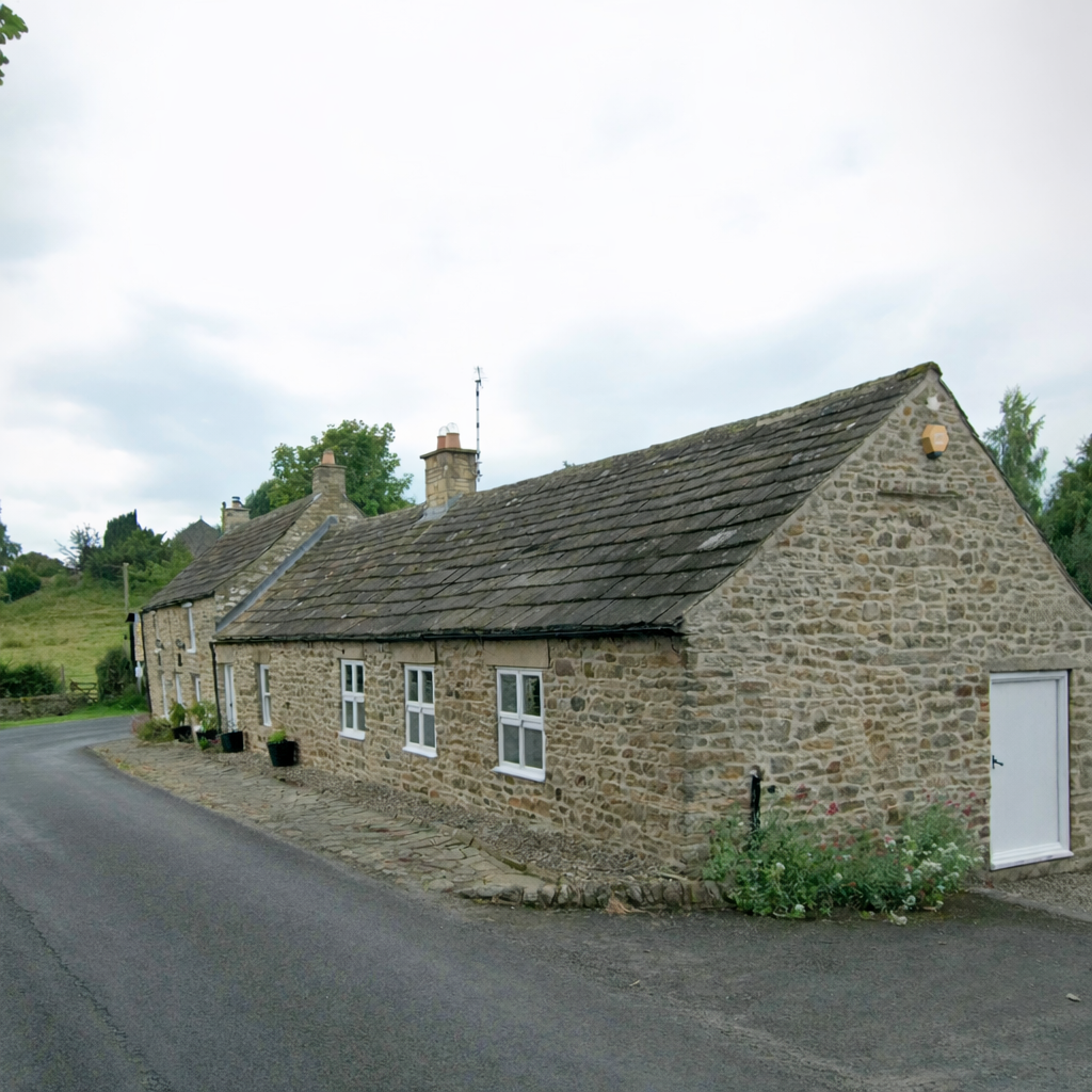 A stone cottage with lime mortar pointing in Northumberland situated along a curved road in a rural area with green landscape and cloudy sky.