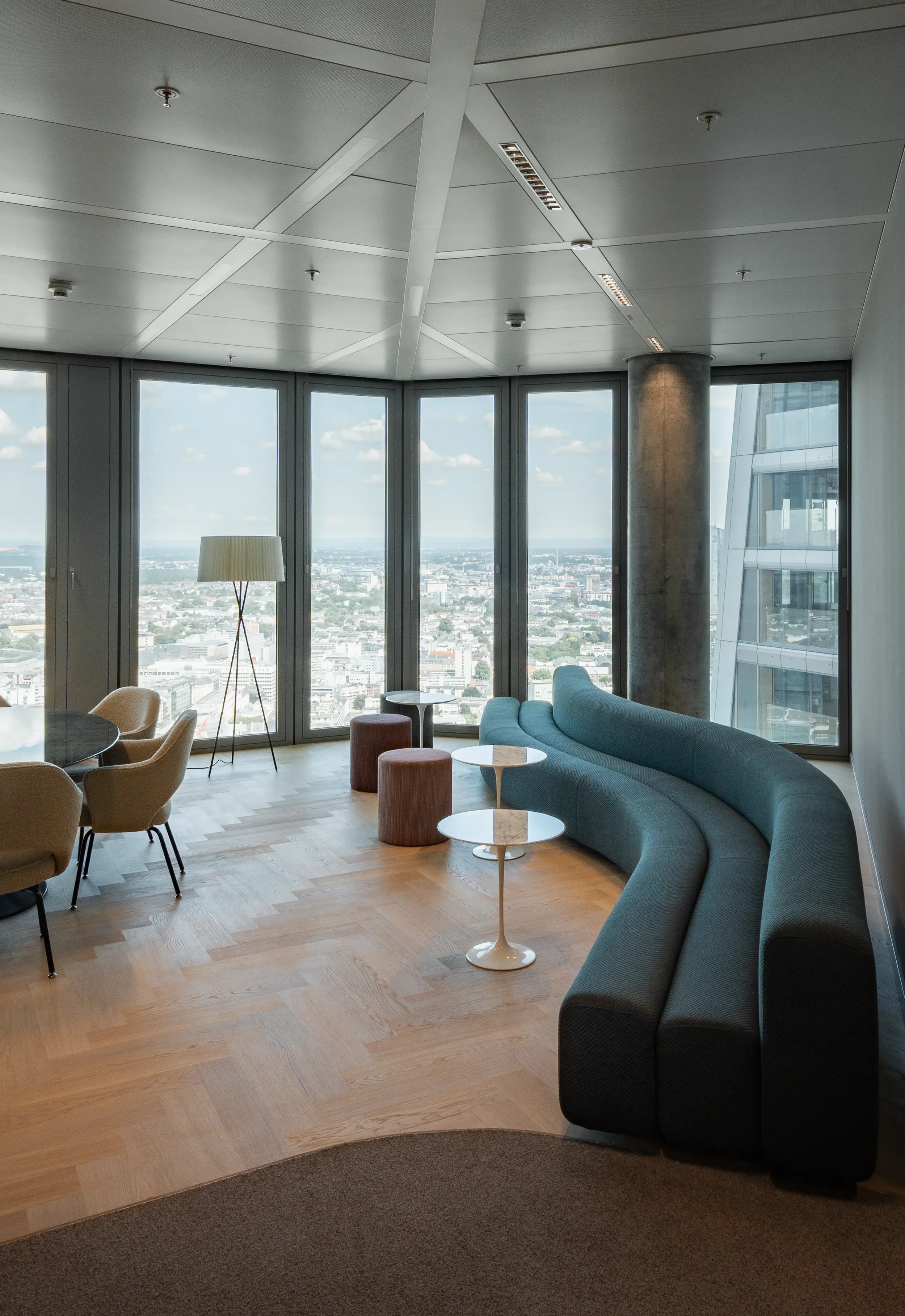 Ein moderner Lounge-Bereich mit großen Fenstern, einer blauen Couch, einem stehenden Stehlampe, kleinen Tischen und einigen Stühlen, Blick auf die Stadt im Hintergrund.