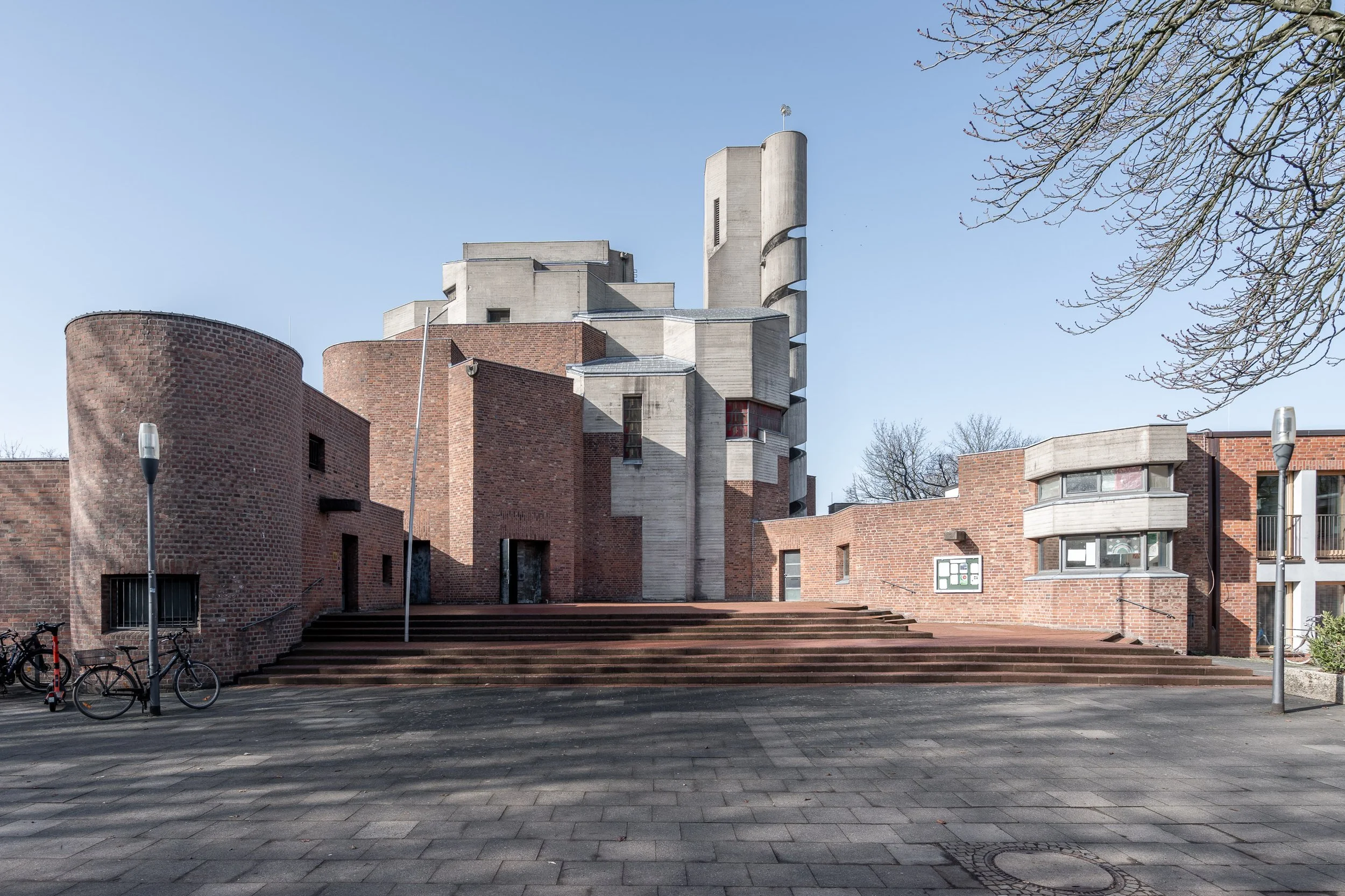 Modernes Gebäude mit roten Ziegelwänden, weißen Betonstrukturen und mehreren rundlichen Elementen, umgeben von einer gepflasterten Fläche und Bäumen mit Blättern