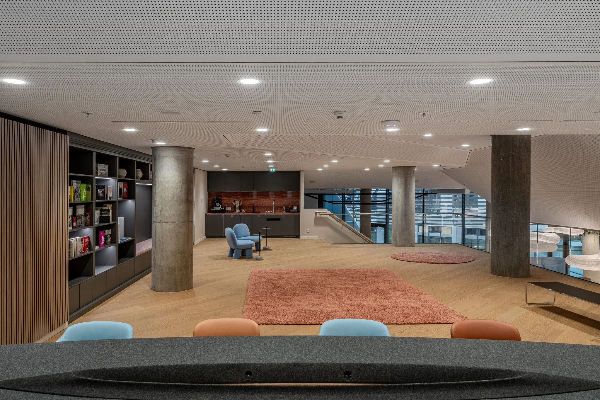 Modernes, offenes Wohnzimmer mit Holzboden, großen Fenstern, einer kleinen Bar mit Küchenzeile, zwei blue Sessel, einem abgerundeten Sitz, Teppichen und Bücherregal.