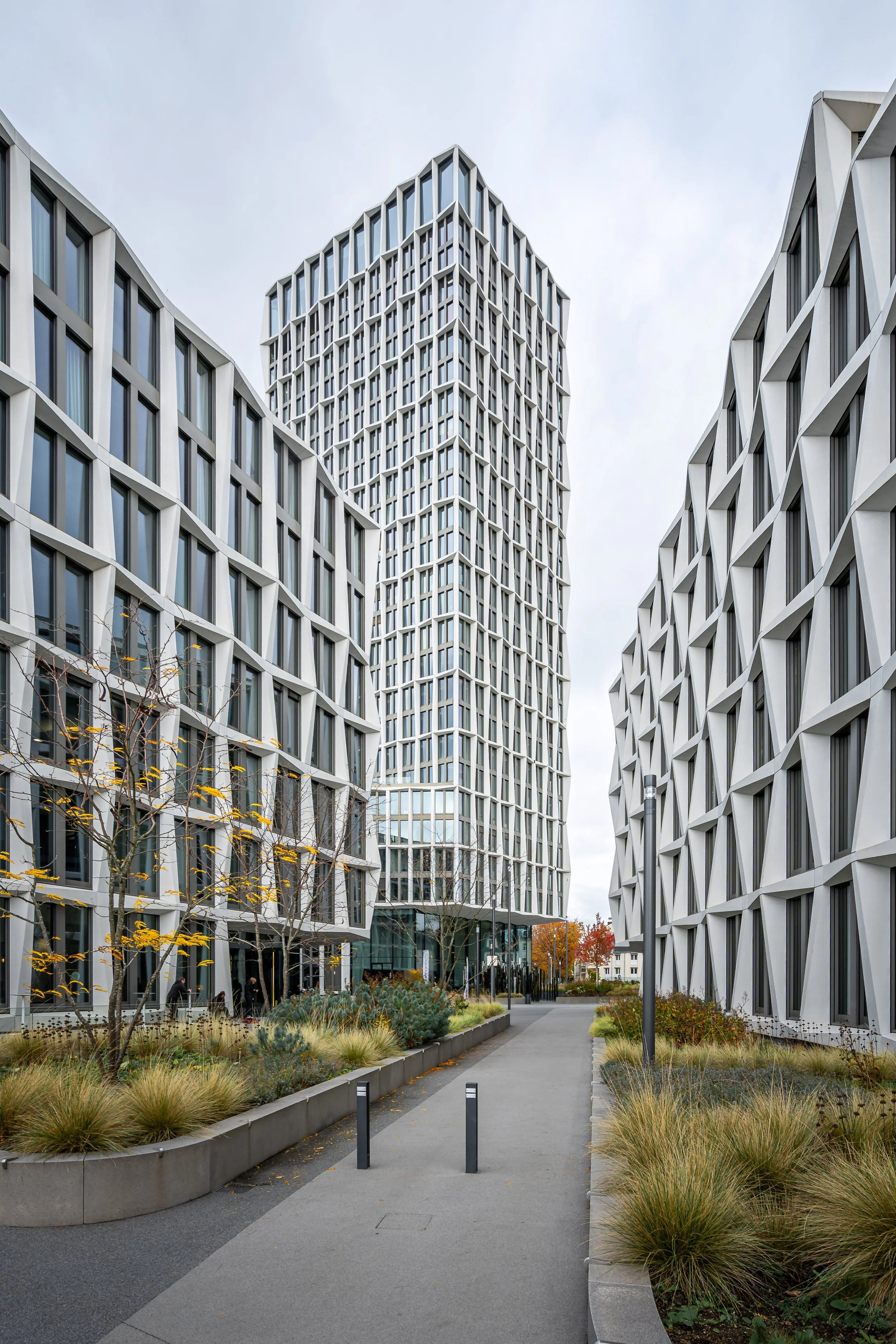 Moderne Bürogebäude mit geometrischer Fassade und einem gepflegten Spazierweg im Vordergrund, umgeben von Bepflanzung und Bäumen, bei bewölktem Himmel.
