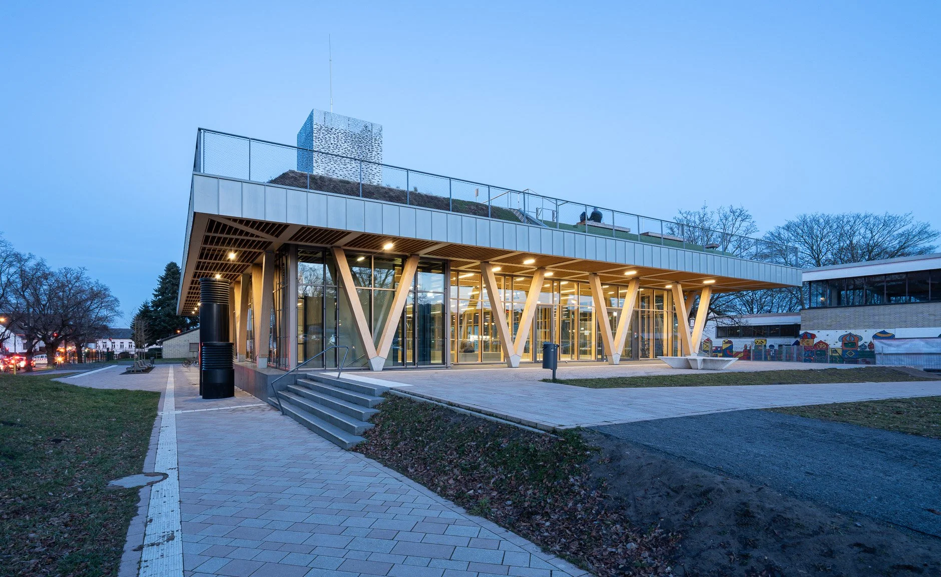 Modernes zweistöckiges Gebäude mit großen Glasfenstern und Holzstützen, umgeben von Bäumen während der Abenddämmerung