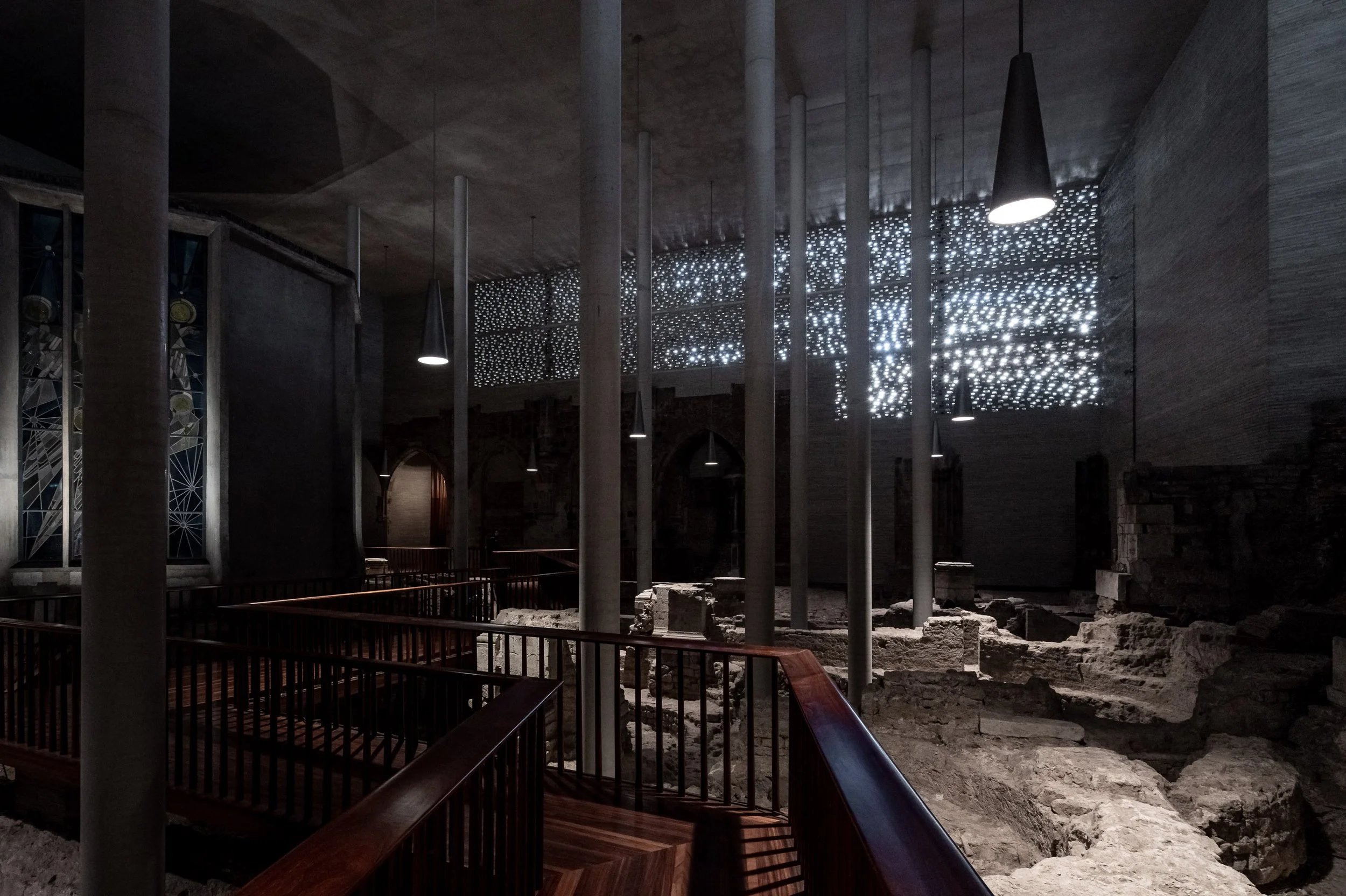 Innenraum einer alten Kirche mit modernen Elementen, große Fenster, Balken und stimmungsvoller Beleuchtung, archäologische Ausgrabungen mit sichtbaren Fundamenten und Steinen im Vordergrund