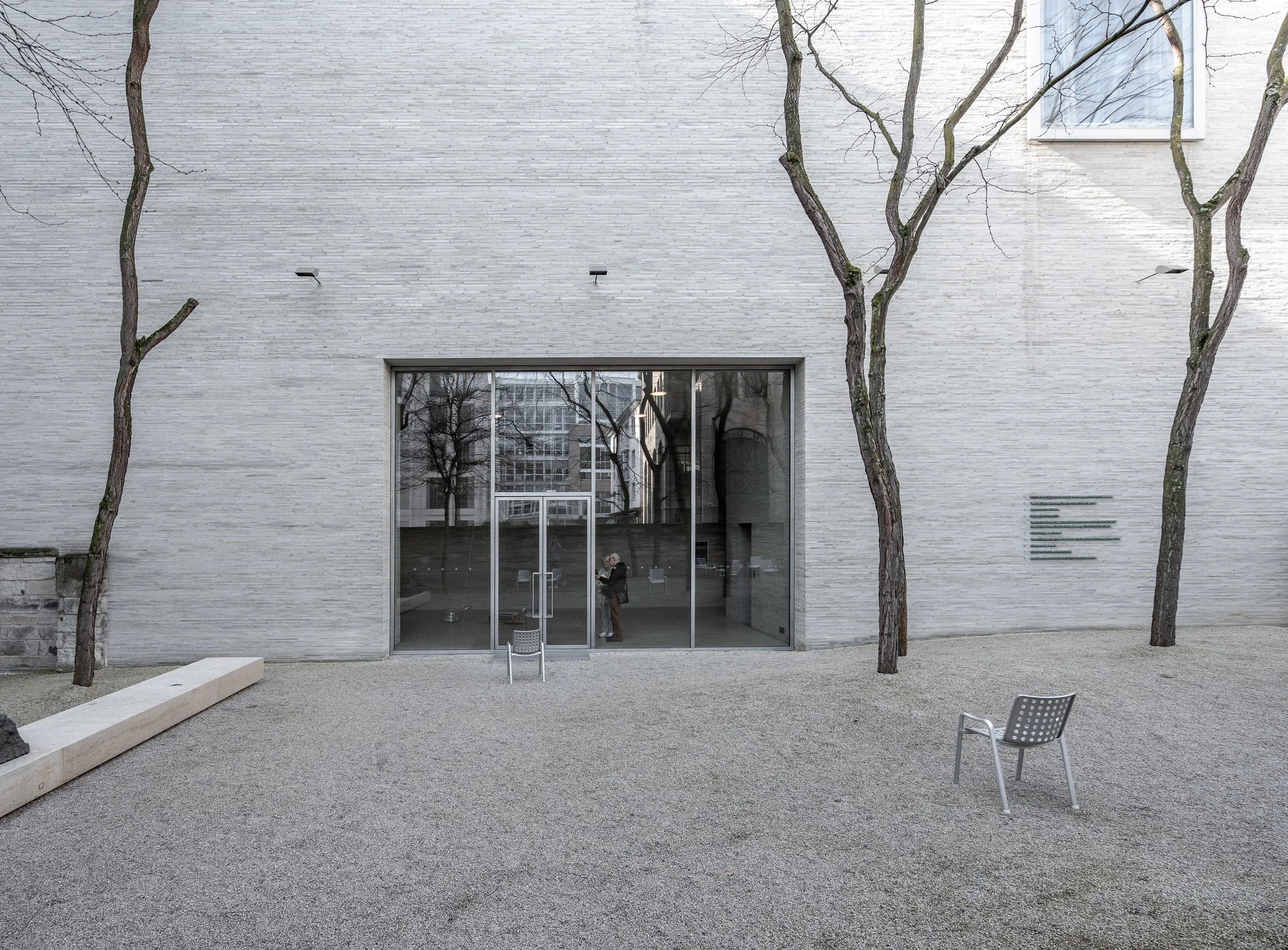 Ein modernes Gebäude mit einer großen Glastür im Eingangsbereich, umgeben von wenigen Bäumen. Es gibt zwei metallene Sitze im Außenbereich, der Boden ist mit Kies bedeckt, und im Hintergrund sind reflektierende Fenster sichtbar.
