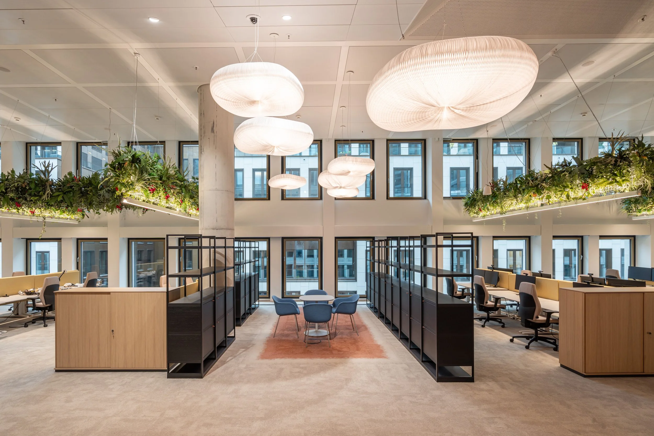 Moderne Büroraum mit großen Fenstern, über dem die Decke mit Hängepflanzen und großen weißen Lampenschirmen dekoriert ist. Es gibt Arbeitsplätze mit Stühlen und Tischen und eine kleine Sitzgruppe in der Mitte auf einem roten Teppich.