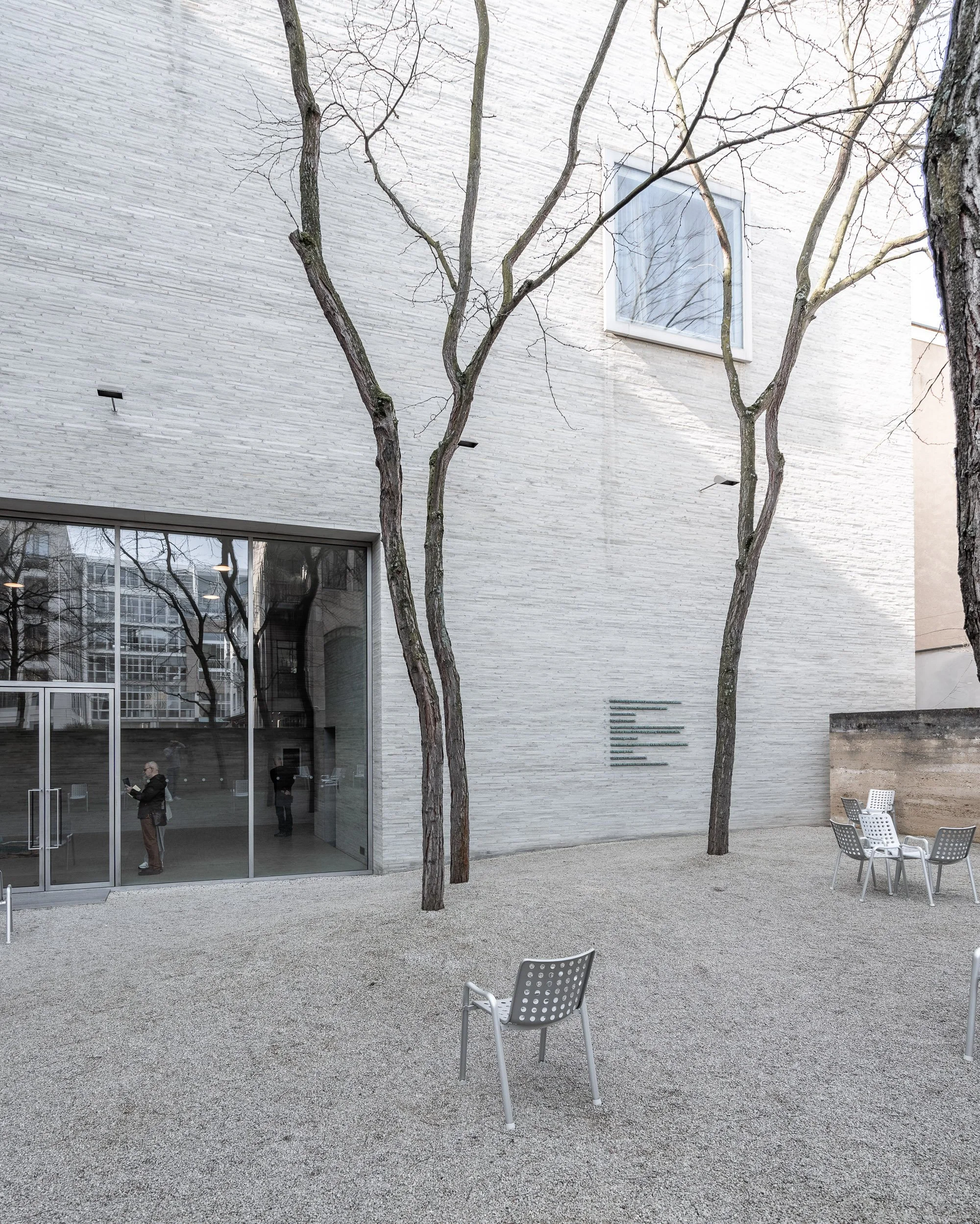 Ein moderner, weißer Gebäudefront mit großen Glasfenstern, vier büschelartige Bäume, ein Gartenbereich mit Sitzgelegenheiten und einem Kiesboden.