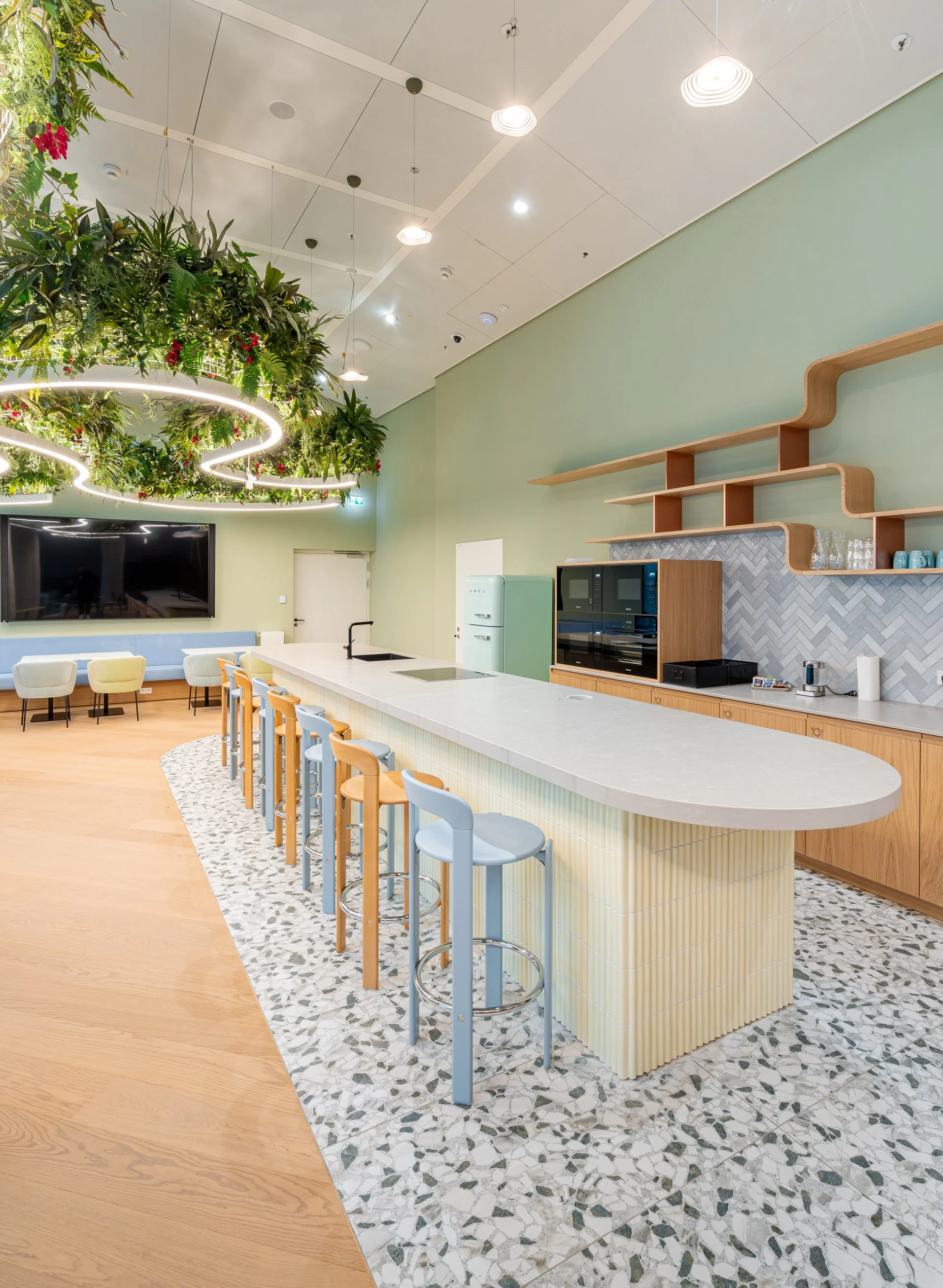 Moderne Cafeteria im FOUR Frankfurt mit Barista-Bereich, großen Fenstern, Pflanzenhängetröge mit Lichteranzug, gemütlichen Sitzgelegenheiten, einem großen Bildschirm, Holzwänden und einem Kaffeeautomaten.