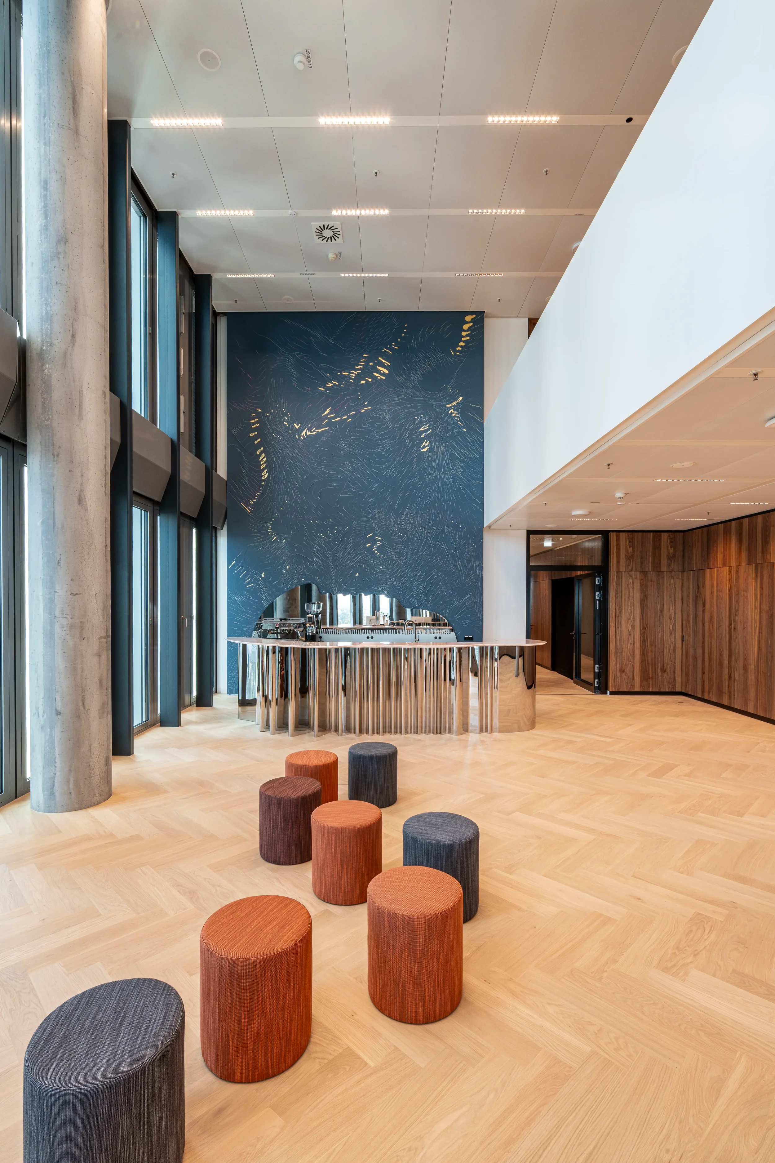 FOUR Frankfurt T4, Moderner Innenraum mit Holzparkett, bunten Hocker und einer Bar vor einer blauen Wand mit goldener Linienzeichnung.