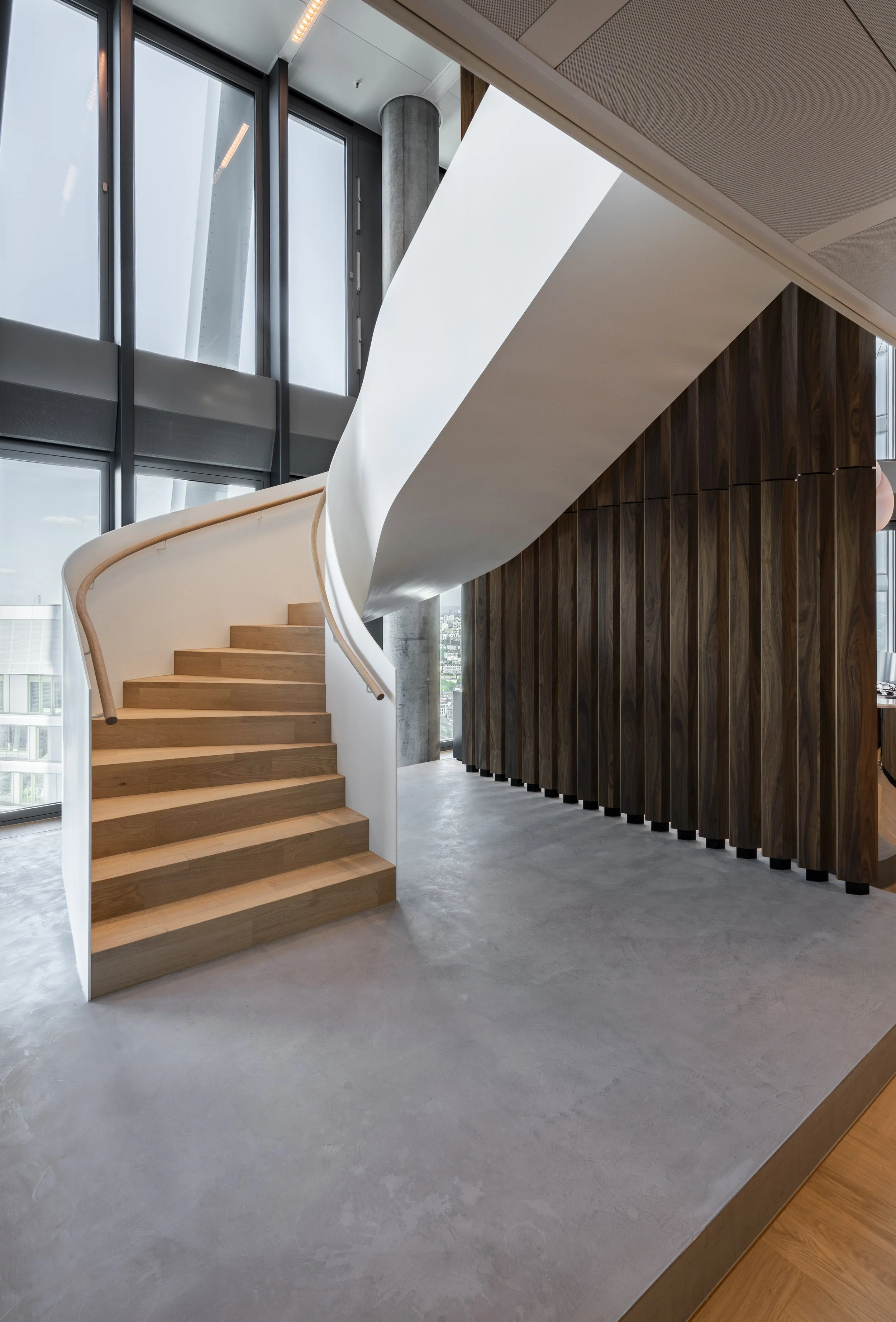 Innenansicht eines modernen Gebäudes mit Holztreppe, weißen Wänden, großen Fenstern und einer dunklen Holzverkleidung.
