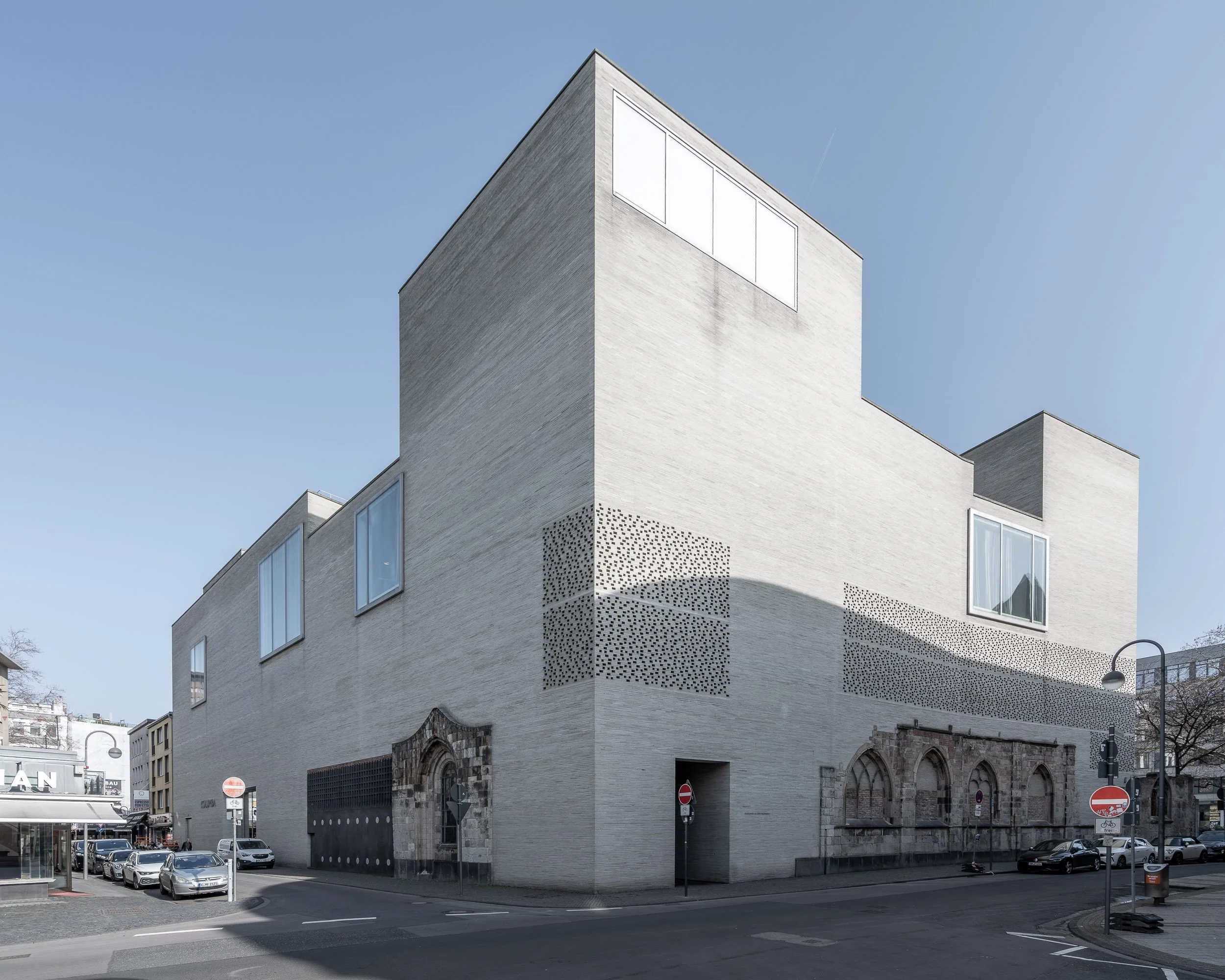 Ein modernes, weißes Gebäude mit großen Fenstern und alten Steinarkaden an der Architektur, auf einer Stadtstraße bei klarem Himmel.