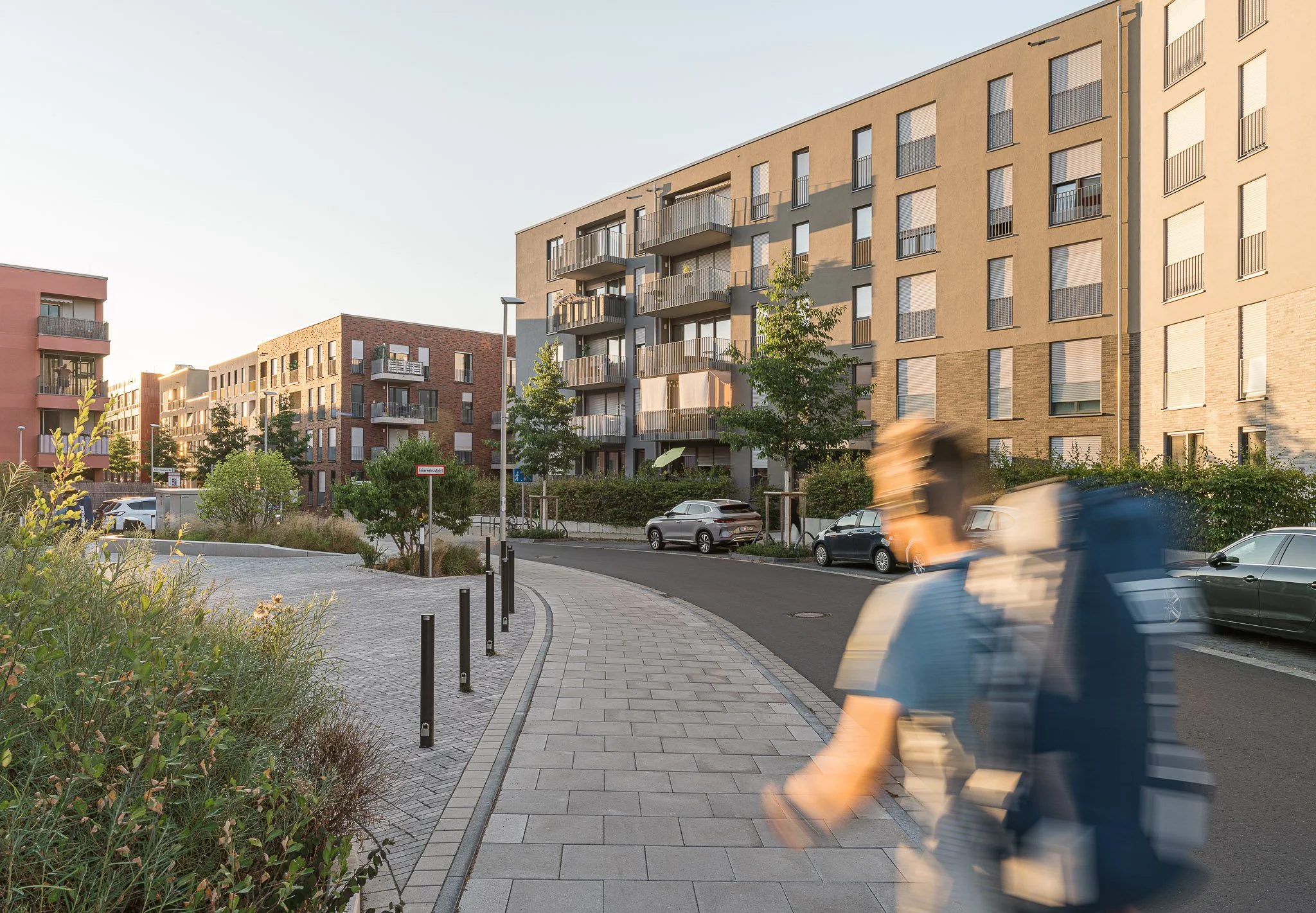 Mehrstöckiges Wohngebäude mit Balkonen und geschlossenem Fenster in einer geplasterten Straße, einige Autos parken und im Vordergrund sind Bäume und Pflanzen, während die Sonne abends auf das Gebäude scheint.
