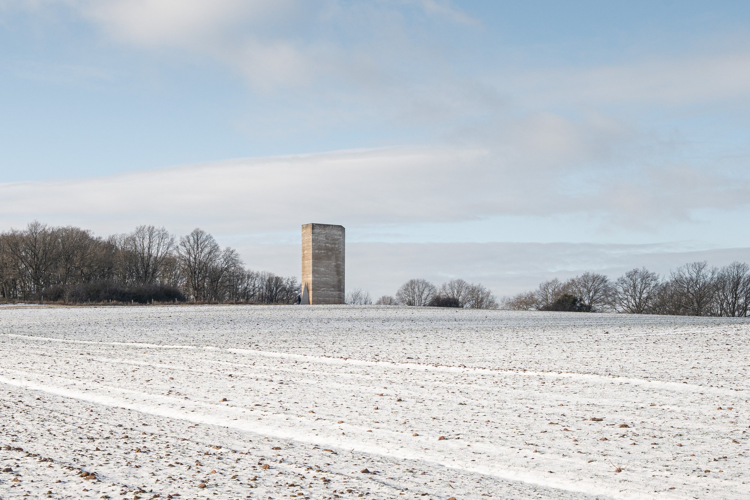 Ein verschneites Feld mit einem einzelnen, rechteckigen Betonbauwerk im Hintergrund und Bäumen am Horizont.