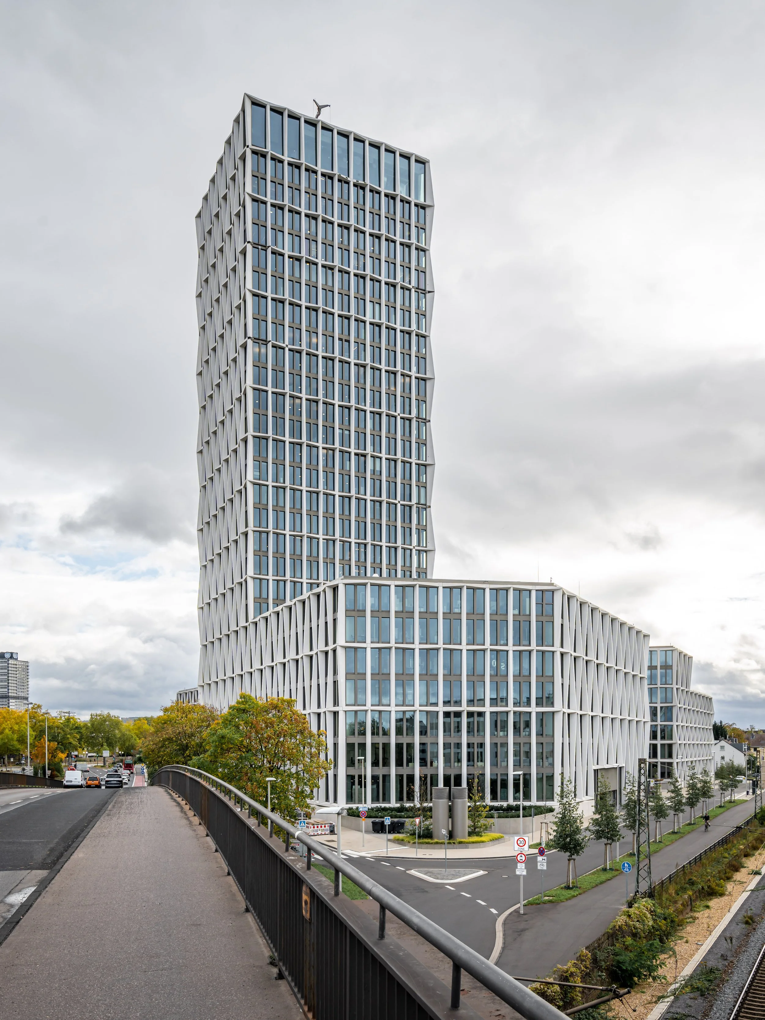 Ein moderner Wolkenkratzer mit gewundener Architektur und großen Fenstern, umgeben von Gehwegen, Bäumen und einer Straße unter einem bewölkten Himmel.