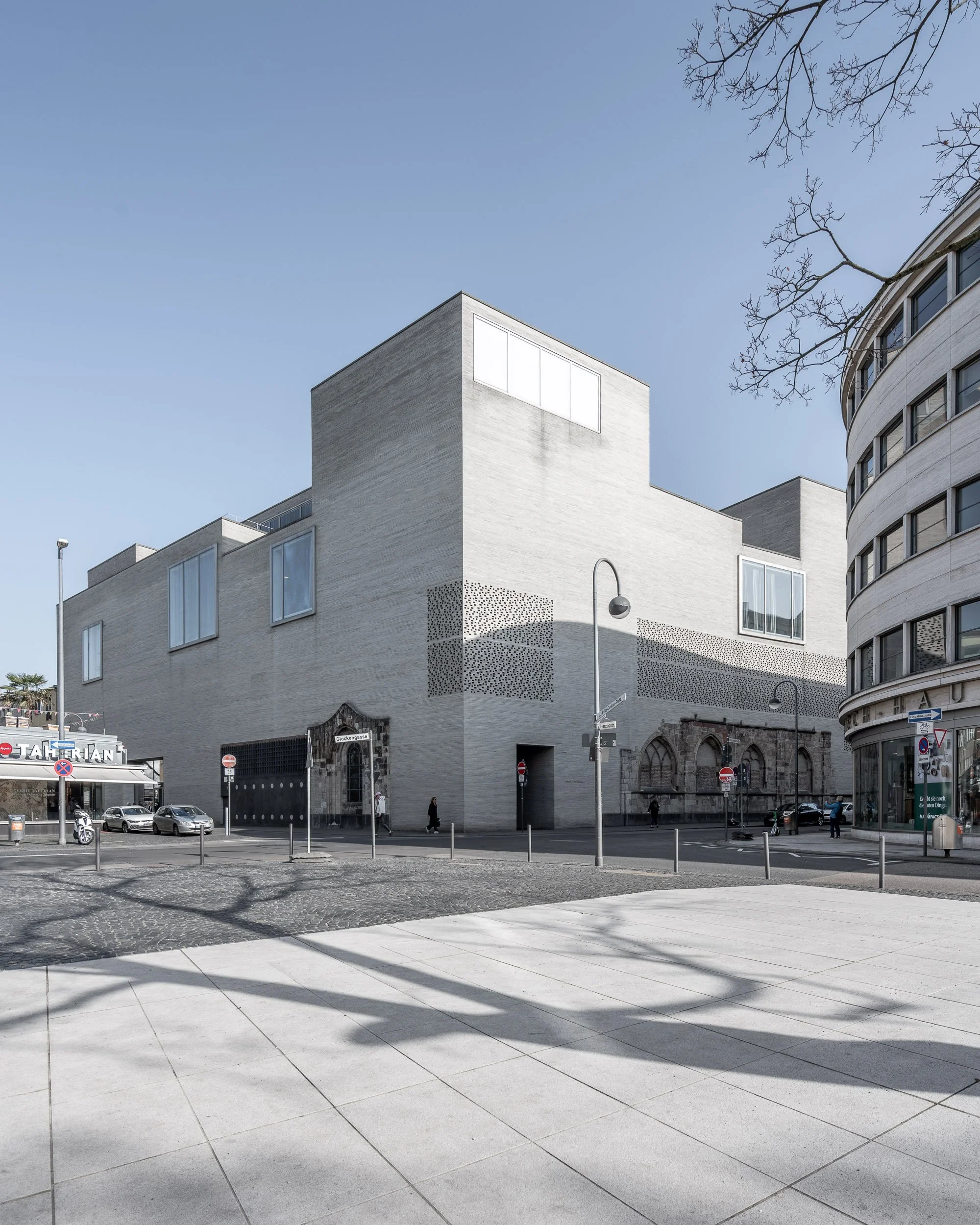 Moderner, weißer Gebäudekomplex mit großen Fenstern an einer städtischen Straße, umgeben von Straßenlaternen, Autos und Passanten, mit einem hellblauen Himmel im Hintergrund.