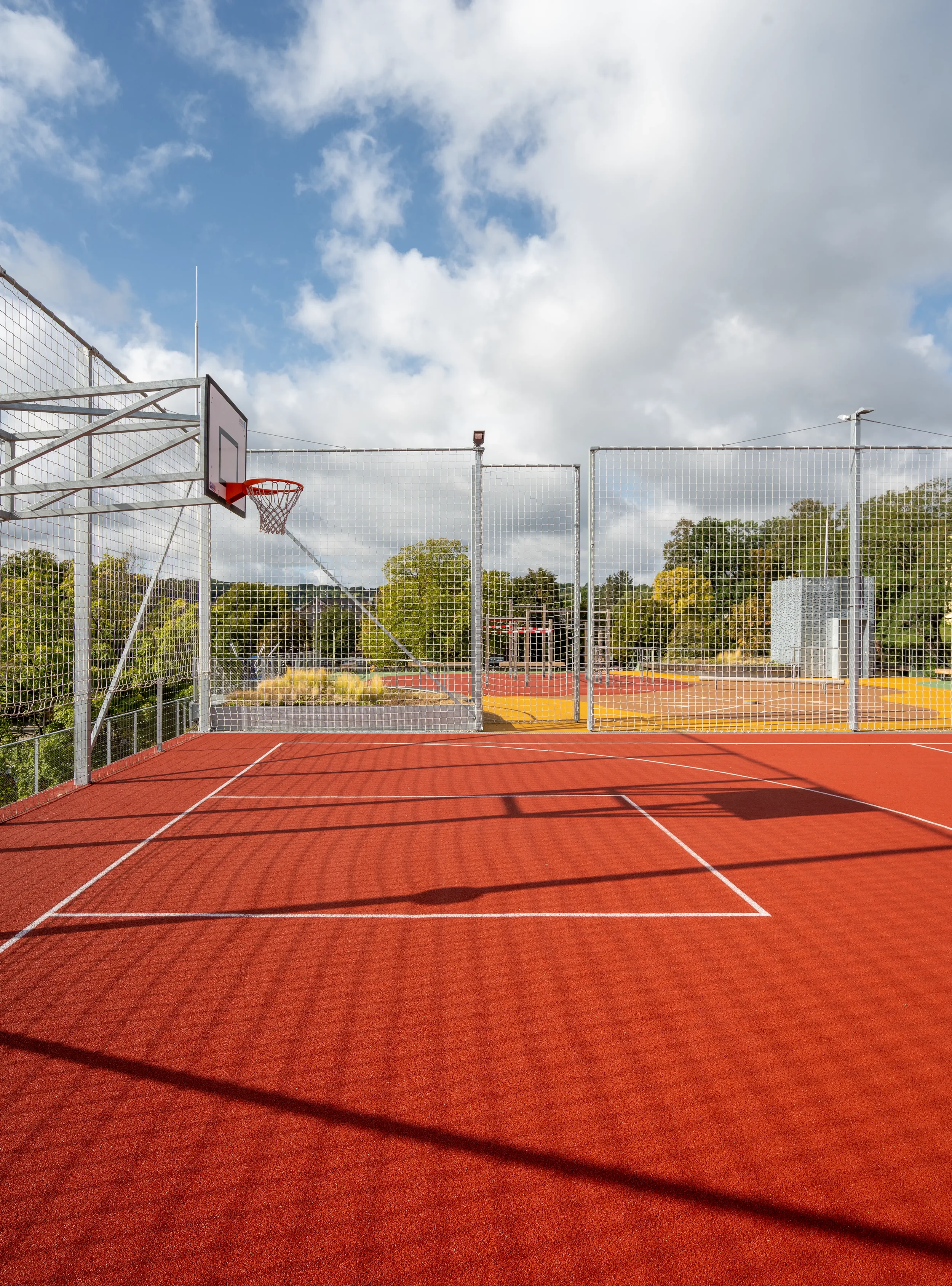 Leerer Outdoor-Basketballplatz unter einem partly wolkigen Himmel, umgeben von einem hohen Gitterzaun.