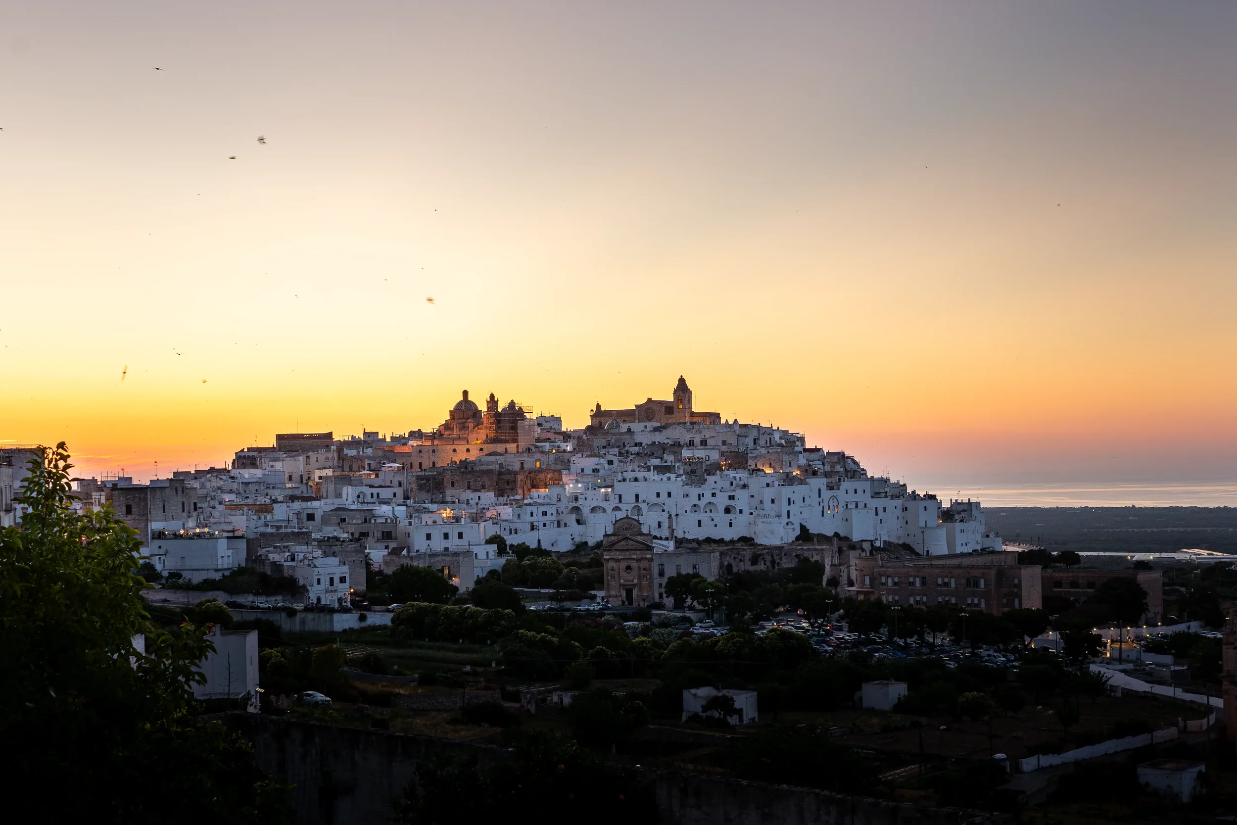 Blick auf die weiße Stadt auf einem Hügel bei Sonnenuntergang, mit einigen Kirchen und Gebäuden, im Hintergrund das Meer.
