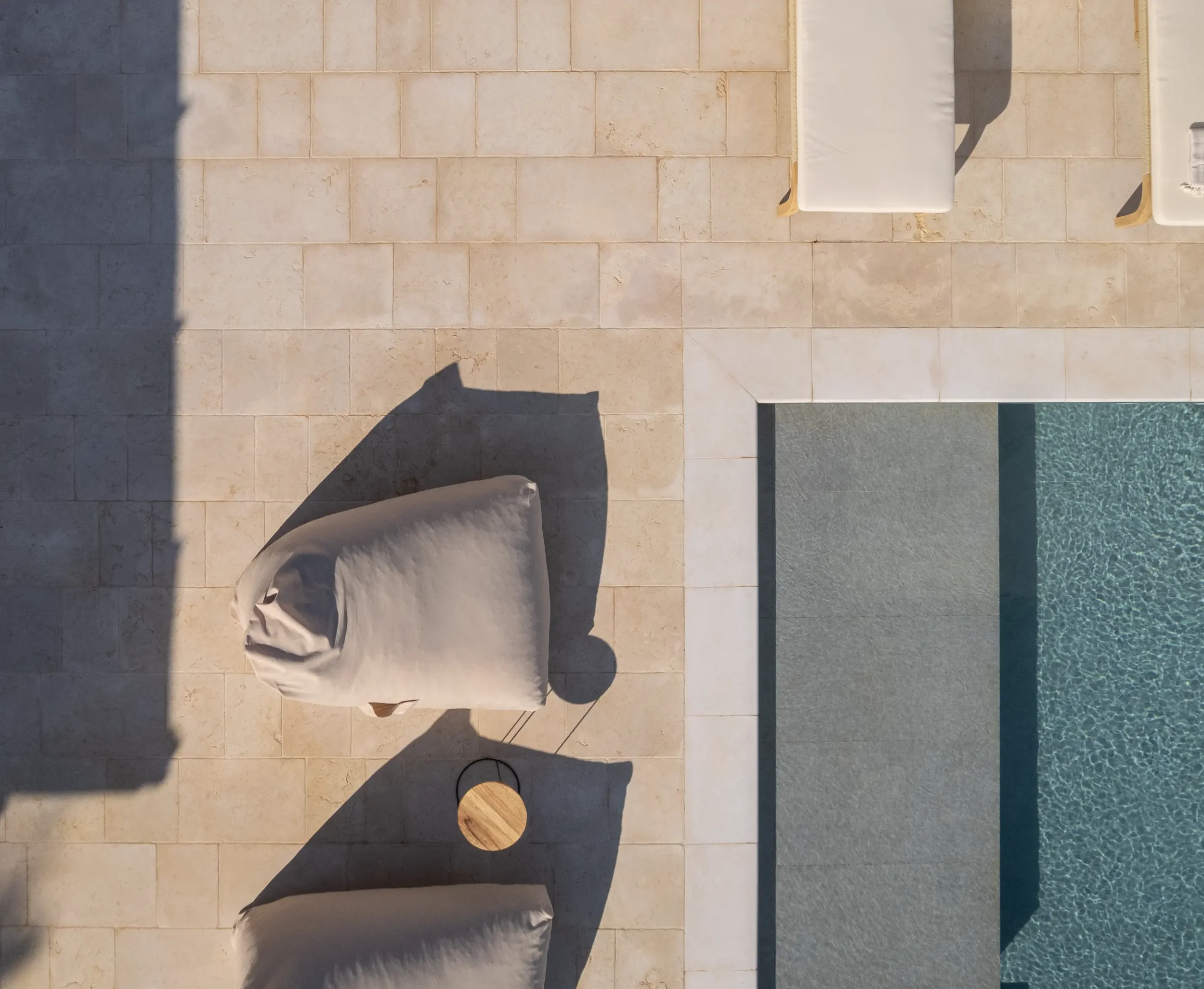 Auf einer Poolterrasse liegen mehrere weiße Liegestühle. Es gibt einen kleinen Tisch und eine Sonne, die Schatten wirft. Ein Pool mit glattem Wasser ist ebenfalls sichtbar.