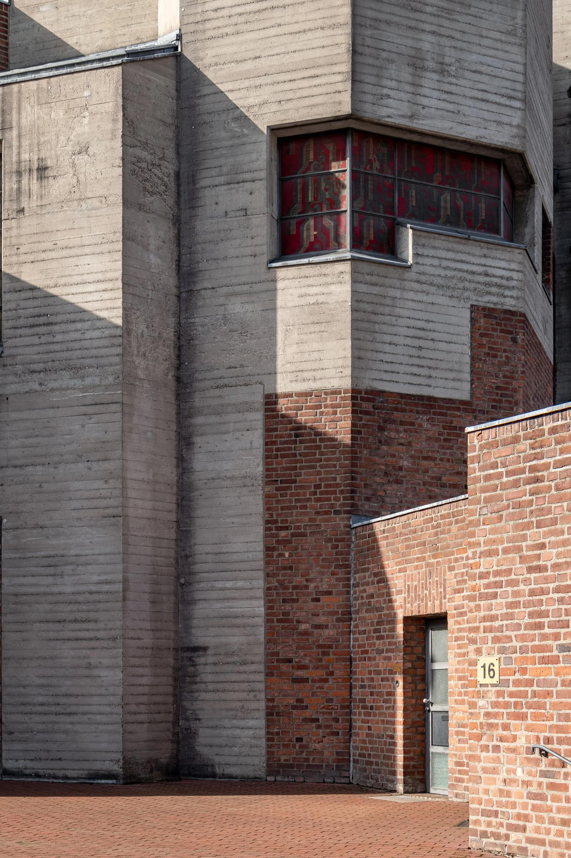 Ansicht eines Gebäudes mit modernen und traditionellen Baumaterialien, darunter Ziegelsteine und Beton, mit einer Tür nummer 16 und einem Fenster mit rotem Muster. Das Gebäude ist bei Sonnenlicht teilweise im Schatten.