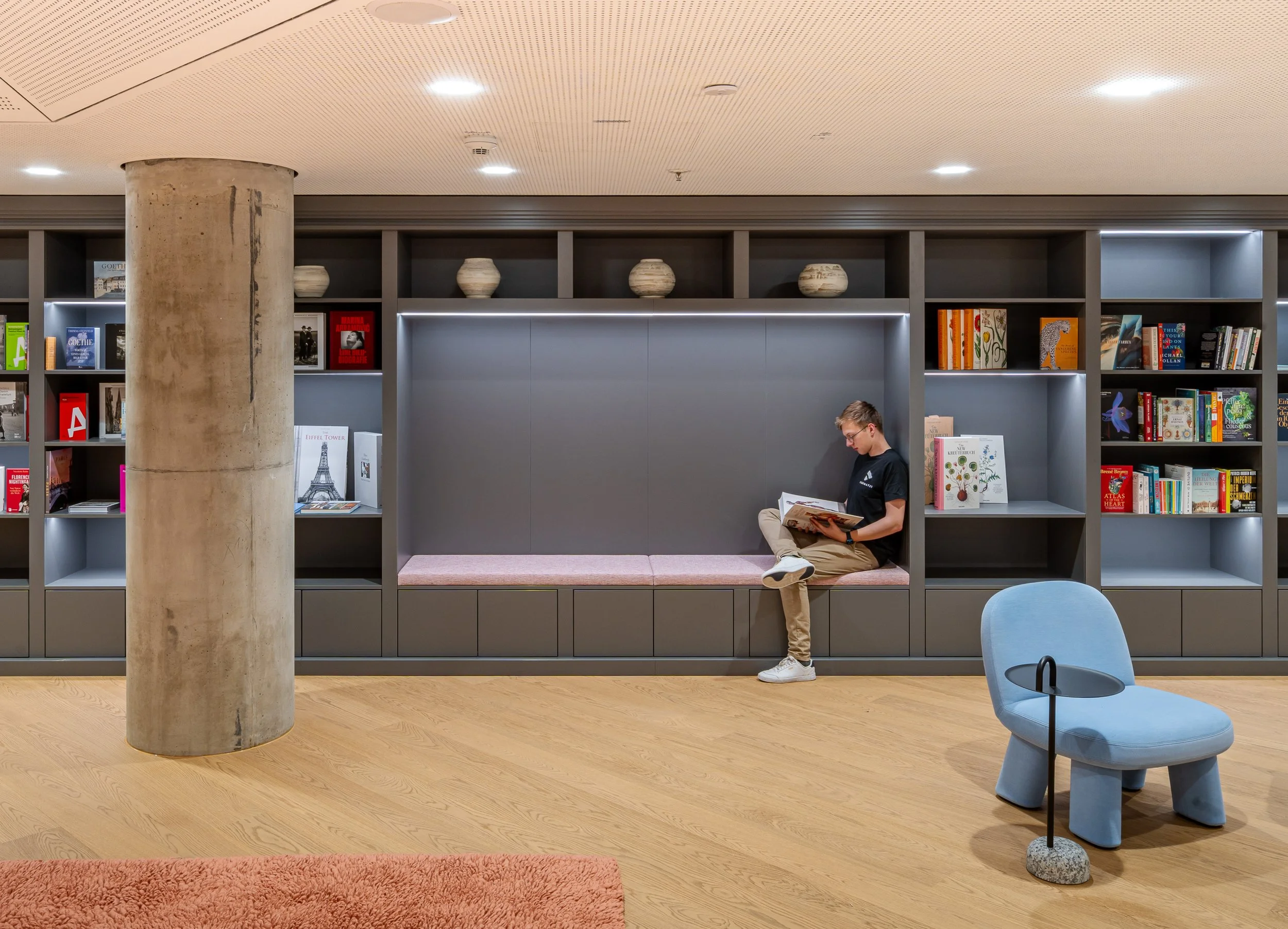 Ein Mann liest in einem modernen Buchladen oder einer Bibliothek, sitzend auf einem Sitzmöbel. Im Hintergrund sind Regale mit Büchern und Dekorvasen, der Raum hat eine angenehme, gut beleuchtete Atmosphäre.