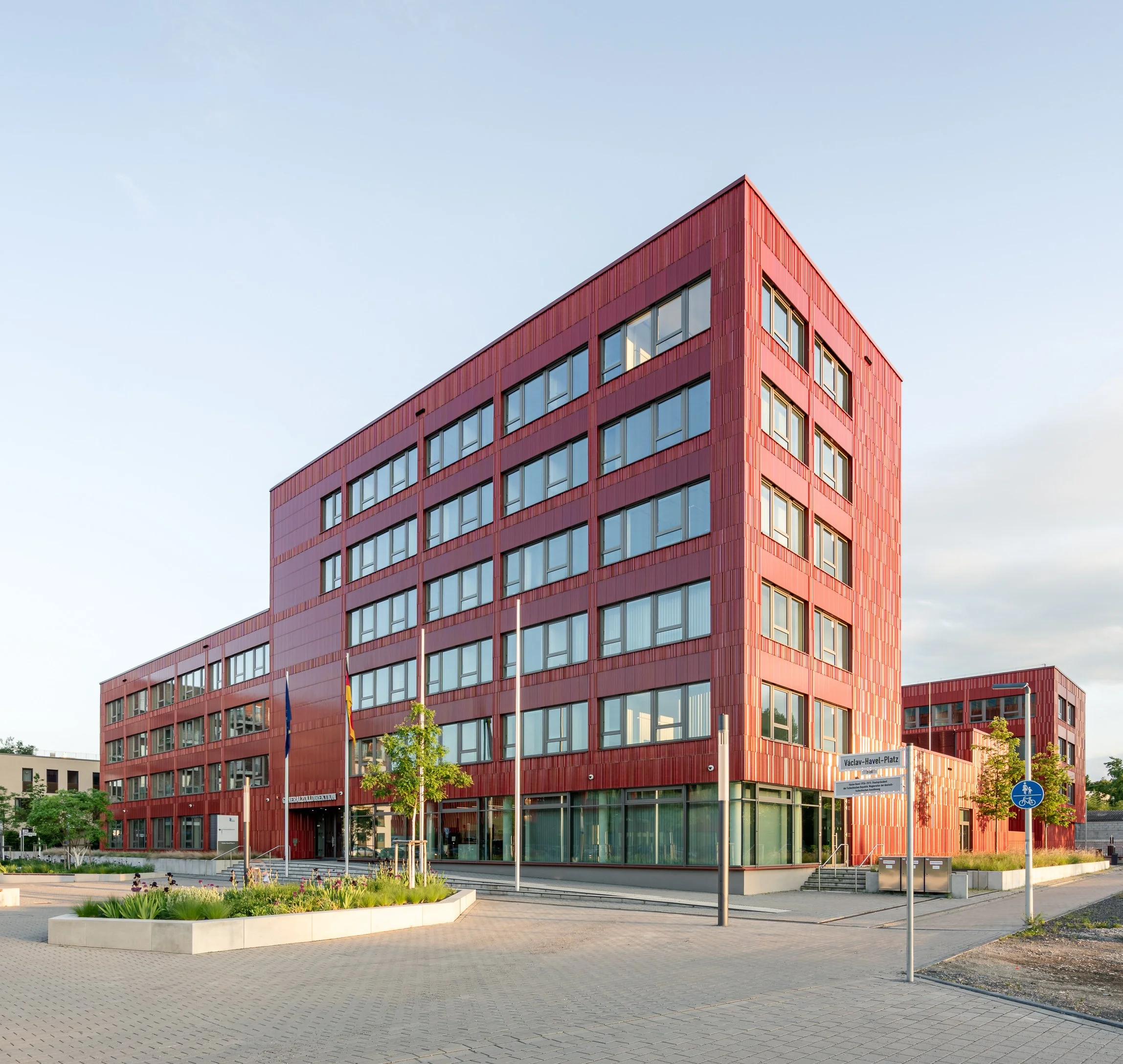 Modern rotes Bürogebäude mit großen Fenstern, Straßenlaternen, Kleinpflaster, Bäume und Blumenbeete davor, Straßenschilder, Himmel im Hintergrund