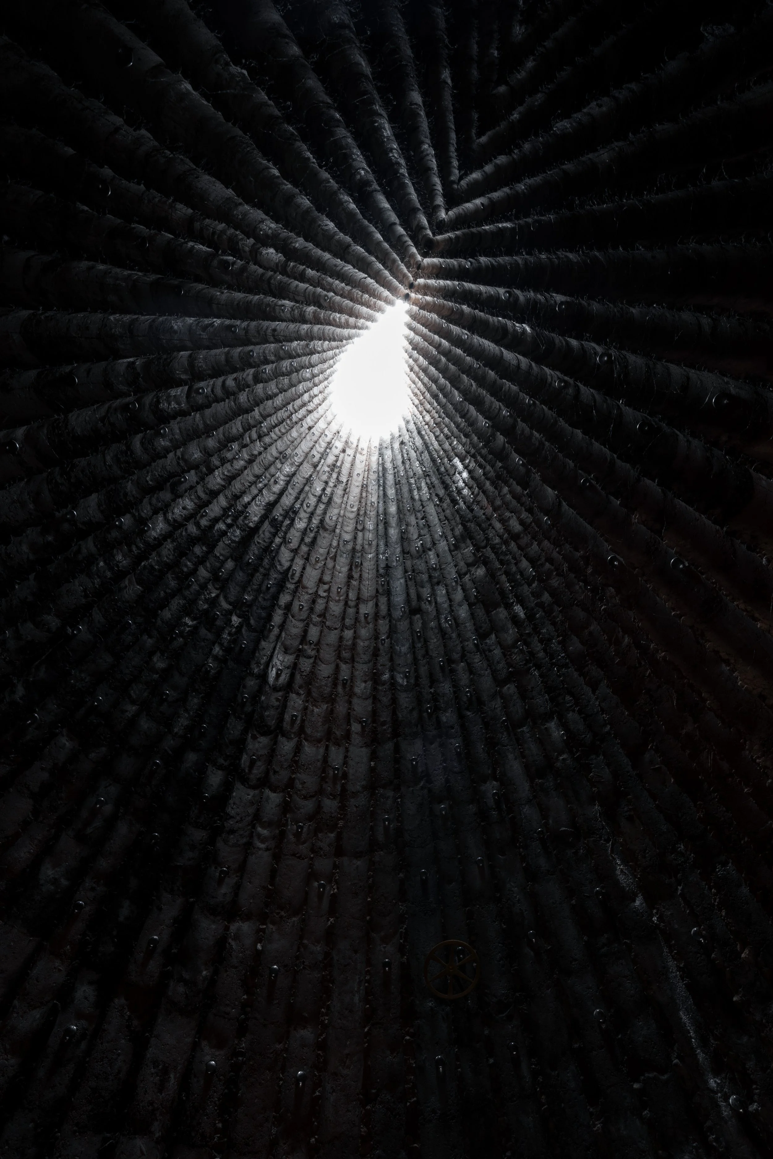 Bruder Klaus Feldkapelle von Peter Zumthor, Innenaufnahme eines Hohlraums mit einer Lichtquelle am Ende, umgeben von schwarzen, runden Gegenständen.