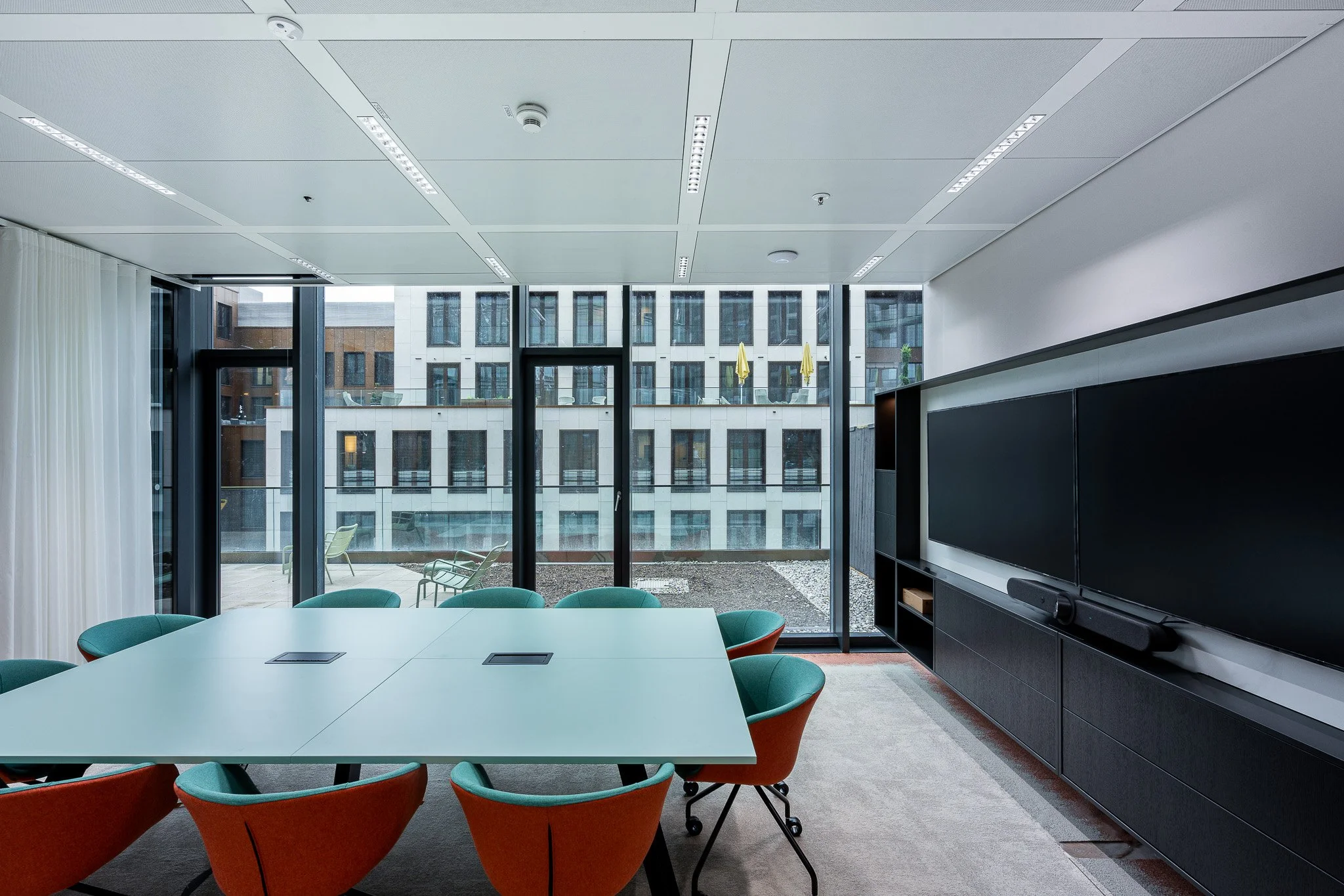 Moderner Konferenzraum mit weißen Tischen, grünen und orangefarbenen Stühlen, großen Fenstern und einem schwarzen Mediencenter mit zwei Bildschirmen.