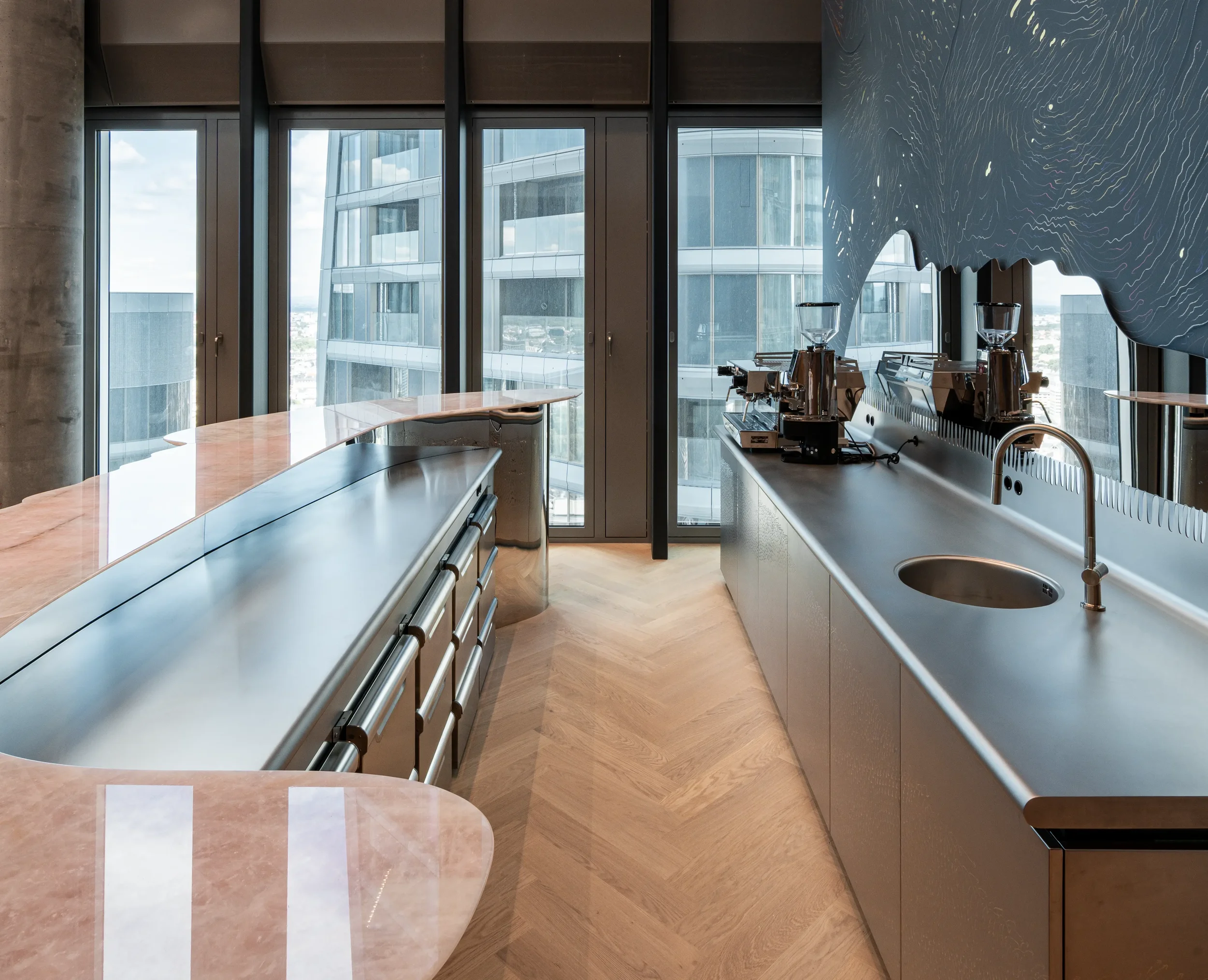 Inneneinrichtung einer modernen Büro-Küche mit großen Fenstern, Küchenarbeitsflächen, Kaffeemaschinen und einer künstlerischen Wandgestaltung.