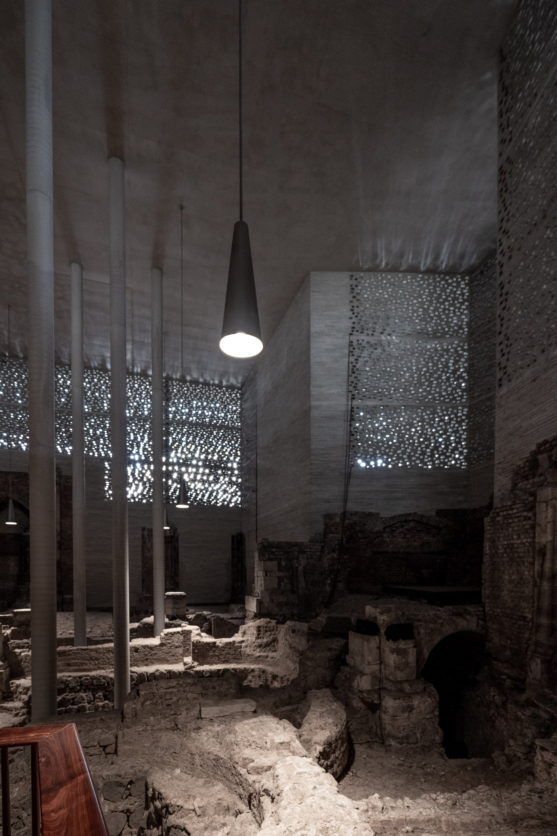 Blick in eine archäologische Ausgrabungsstätte in einem Museum mit moderner Innenarchitektur, inklusive metallene Stützpfeiler, moderne Pendelleuchten, und perforierte metallene Wandpaneele, die Licht durchlassen.