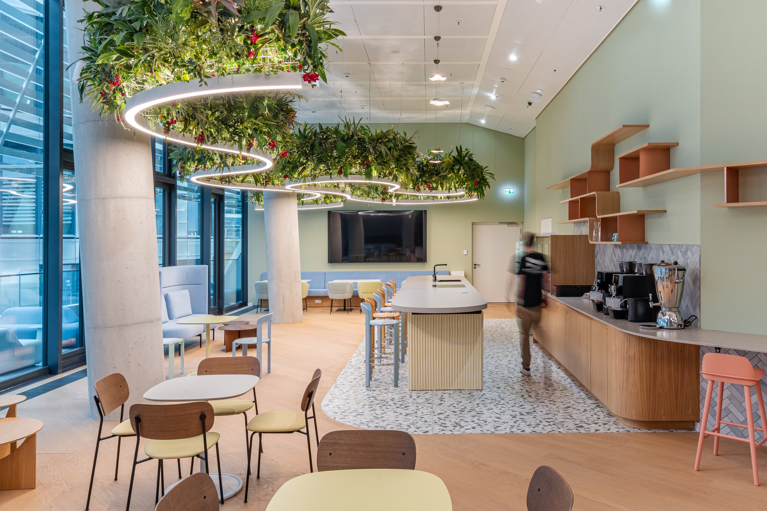 Moderne Cafeteria im FOUR Frankfurt mit Barista-Bereich, großen Fenstern, Pflanzenhängetröge mit Lichteranzug, gemütlichen Sitzgelegenheiten, einem großen Bildschirm, Holzwänden und einem Kaffeeautomaten.