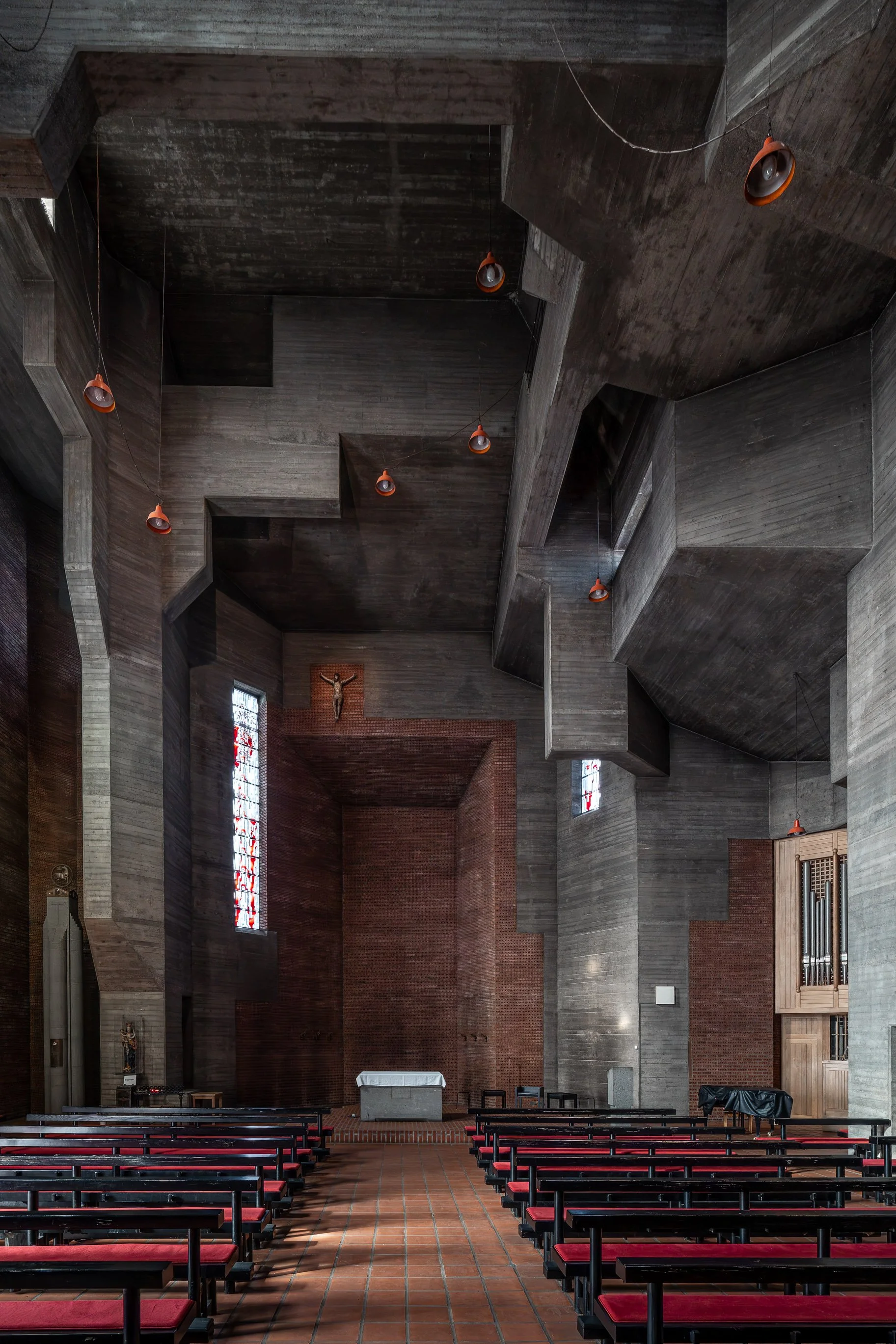 Innenraum einer modernen Kirche mit dunklem Holzdecke, roten Kirchenbänken, einem Altar, einem Kruzifix über dem Altar und bunten Fenster.