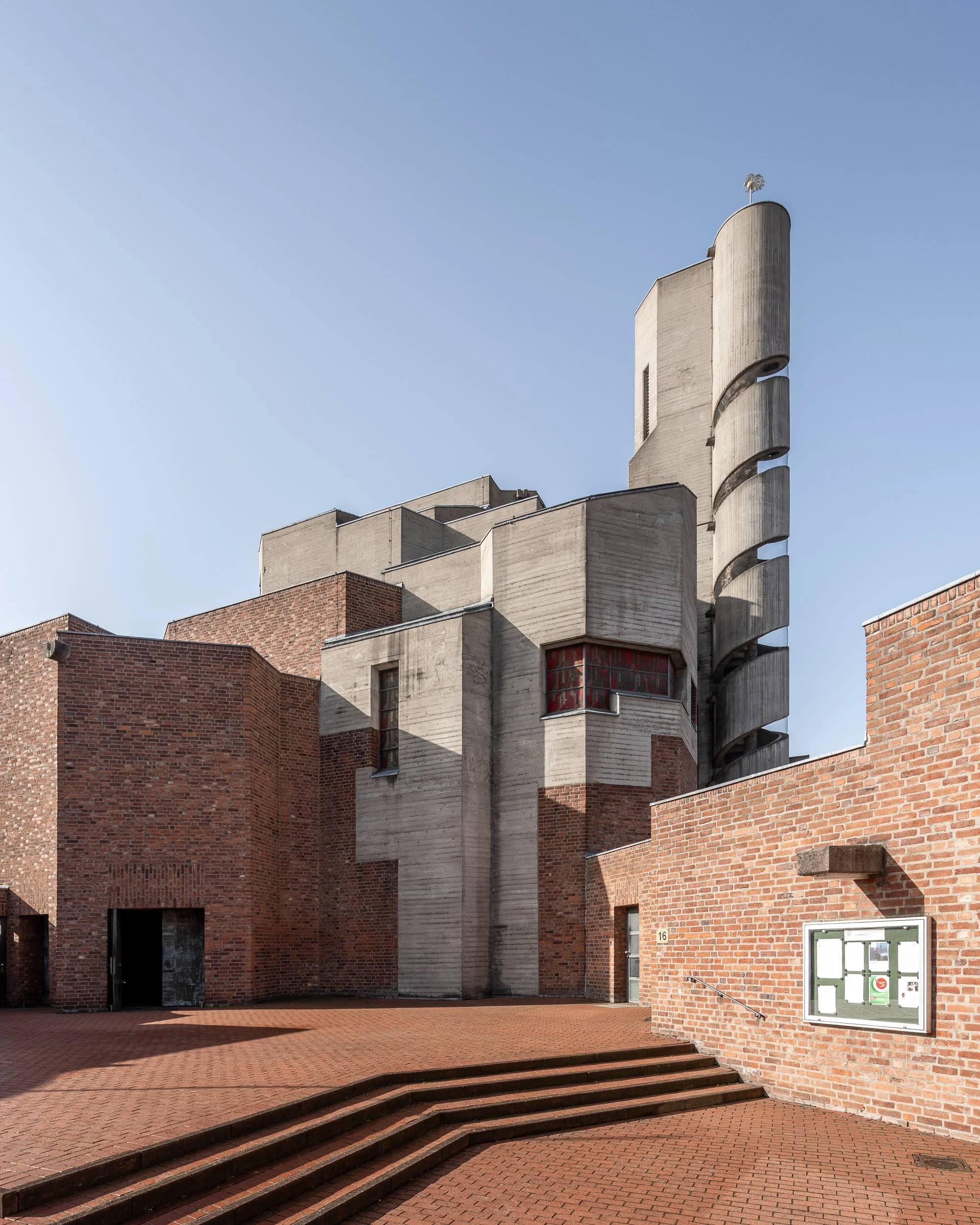 Ein Modernes Gebäude mit einer Mischung aus Backstein und Betonflächen, inklusive einer spiralförmigen Treppe an der Fassade, vor einem blauen Himmel.