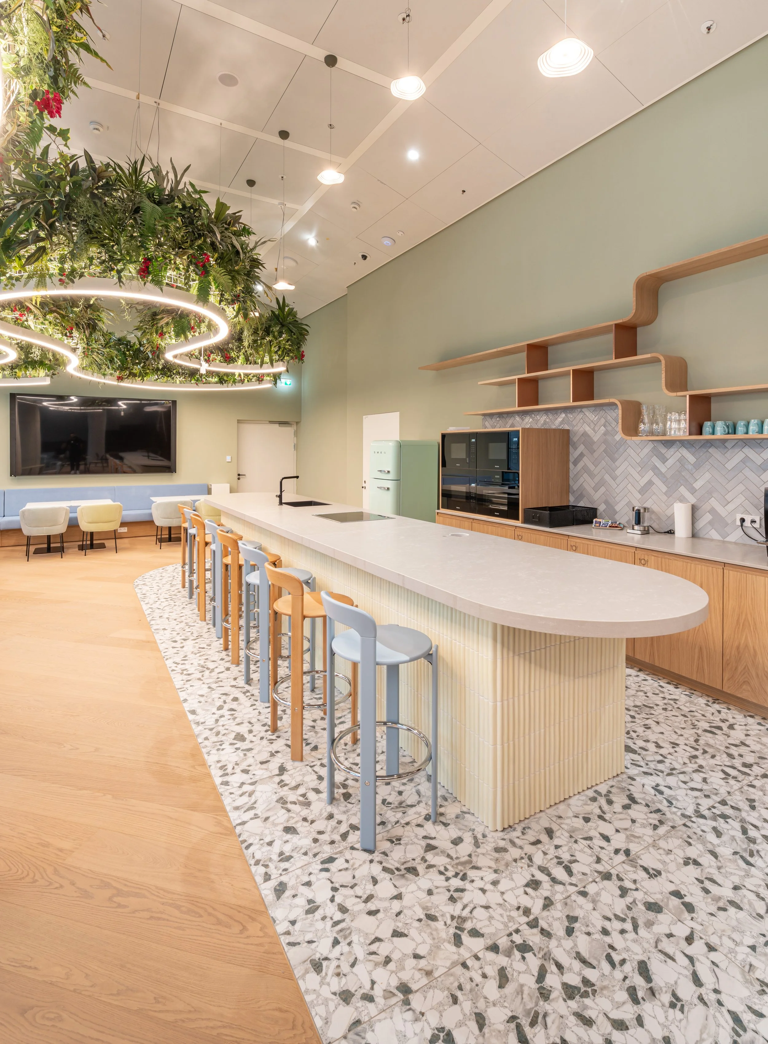 Moderner Gemeinschaftsraum mit Küchenbereich, Theke und Sitzgelegenheiten, große hängende Pflanzen, leuchtende Deckenleuchten, TV an der Wand, helle Farbtöne und gemütliches Ambiente.