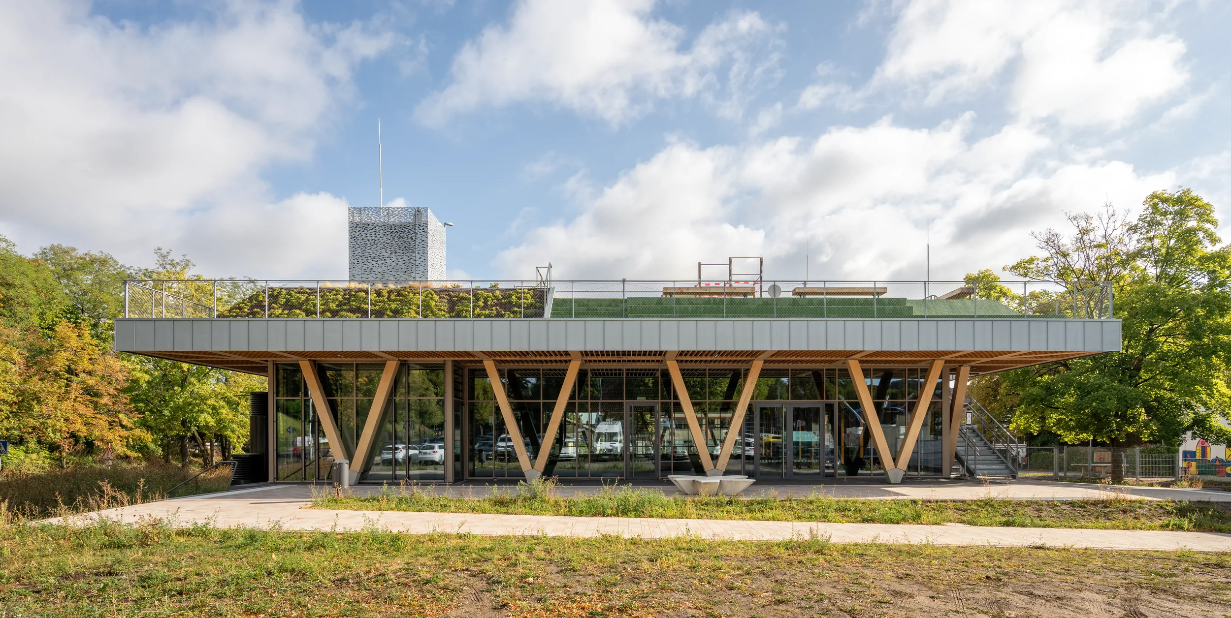 Architekturfotografie Kultur- und Sporthalle in Alfter, Modernes zweistöckiges Gebäude mit großen Glasfenstern und Holzstützen, umgeben von Bäumen und einem gepflegten Außenbereich.
