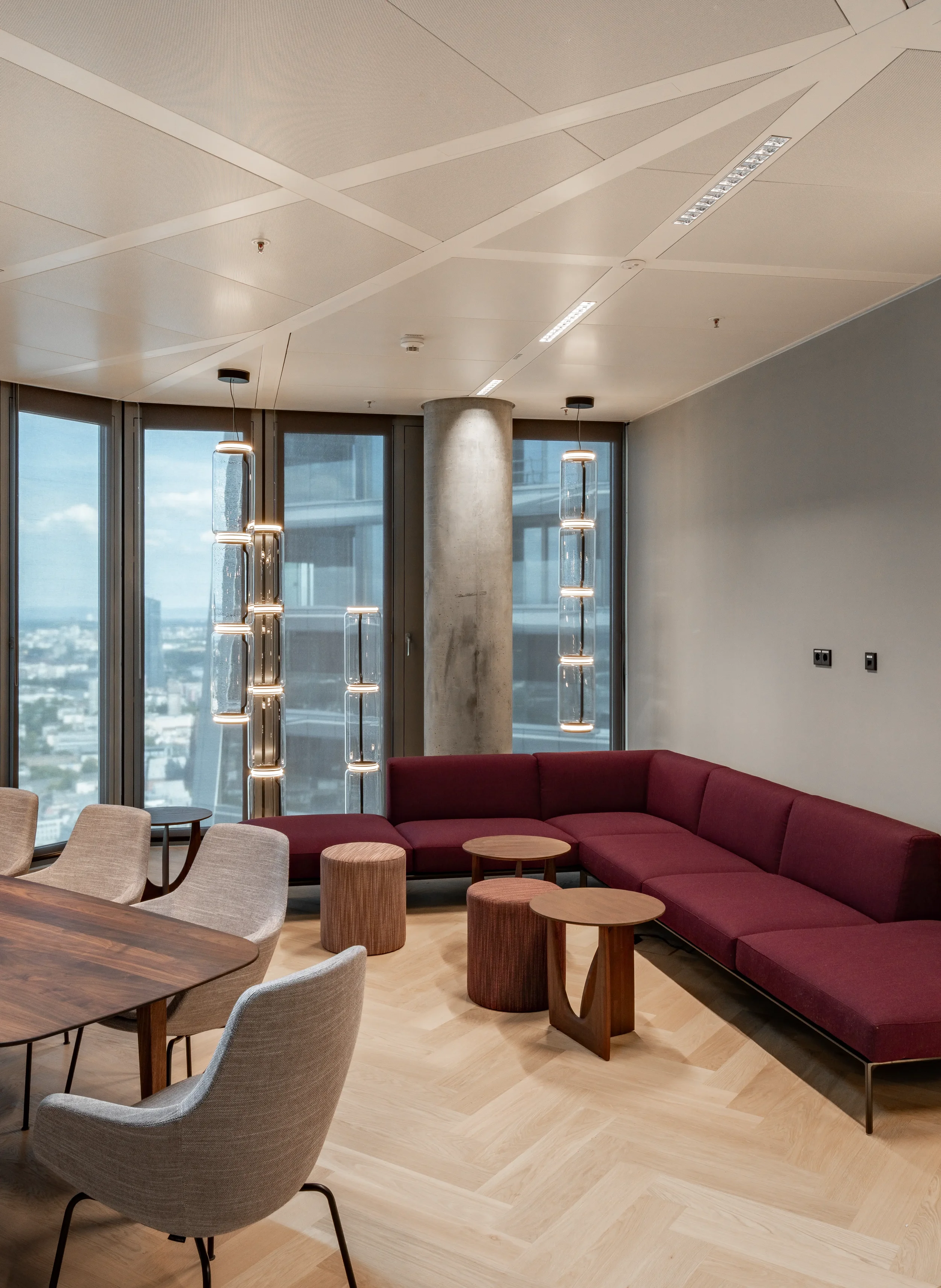 Moderner Aufenthaltsraum mit rotem Sofa, Holztischen, beigen Sesseln, einer großen Fensterfront mit Stadtblick, stilvollen Deckeneinbauten und ungewöhnlichen Stehlampen.