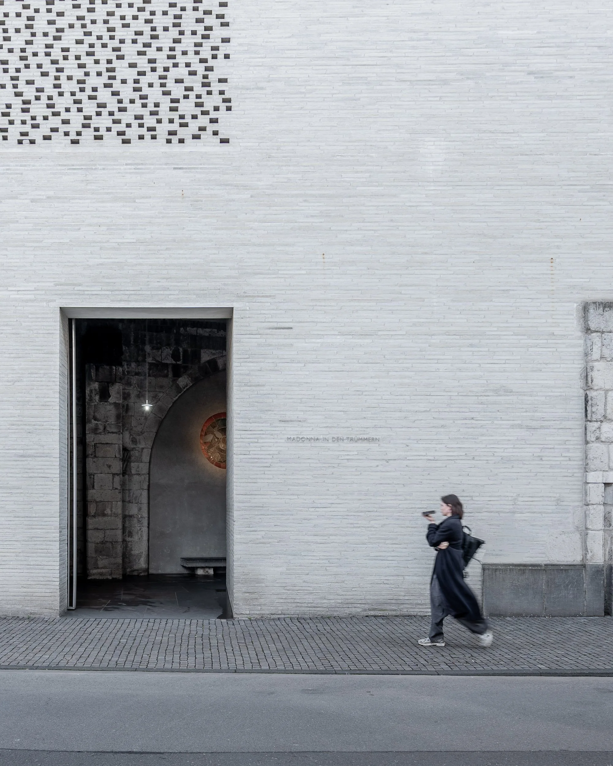 Eine Person läuft auf dem Bürgersteig an einer weißen Ziegelwand und schaut auf ihr Smartphone.