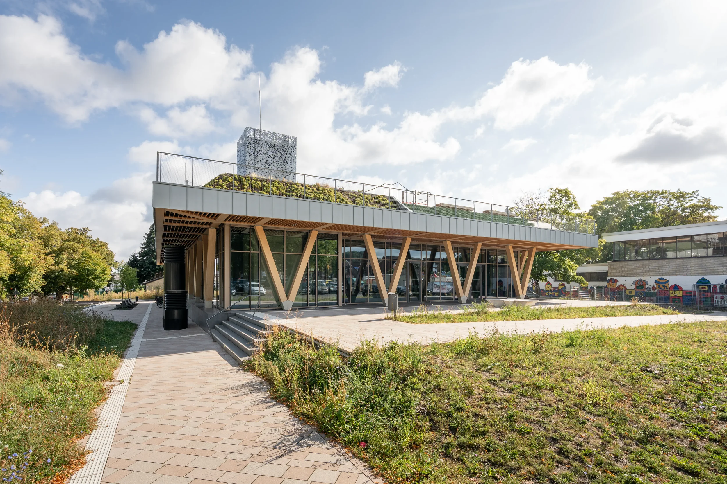Modernes Gebäude mit Holzstützen, Glasfassade und begrünter Dachterrasse, im Freien bei Sonnenschein, mit Himmel und Wolken im Hintergrund.