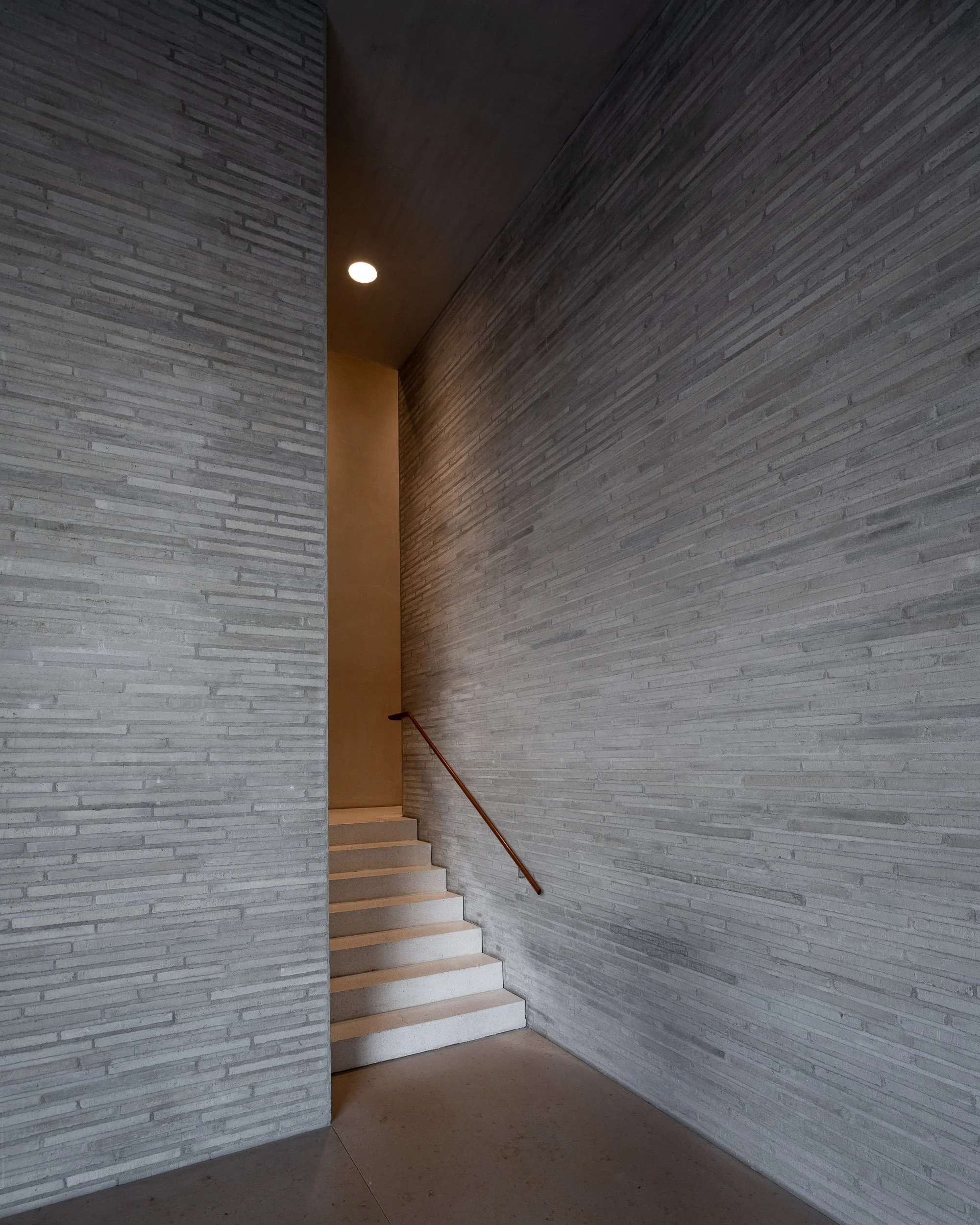 Innenaufnahme eines modernen Treppenhauses mit weißen Stufen, grauen Wandfliesen und einer Holzhandlauf