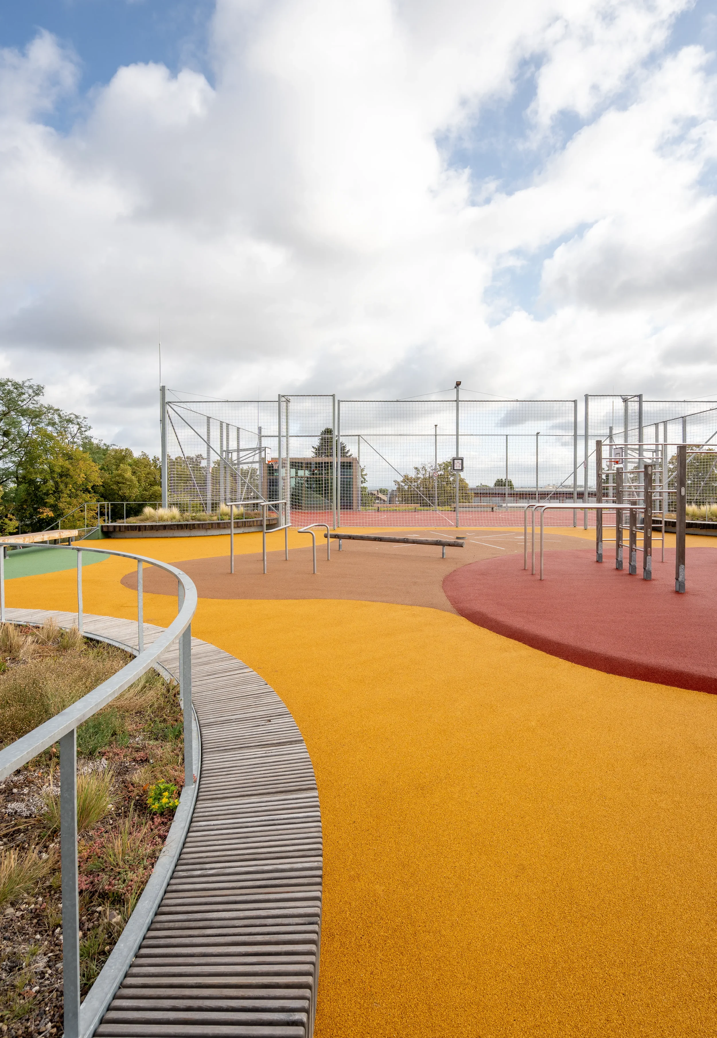 Bühne auf einem Spielplatz mit bunt gestrichenen Bodenflächen und einem hohen Zaun im Hintergrund, bei bewölktem Himmel.
