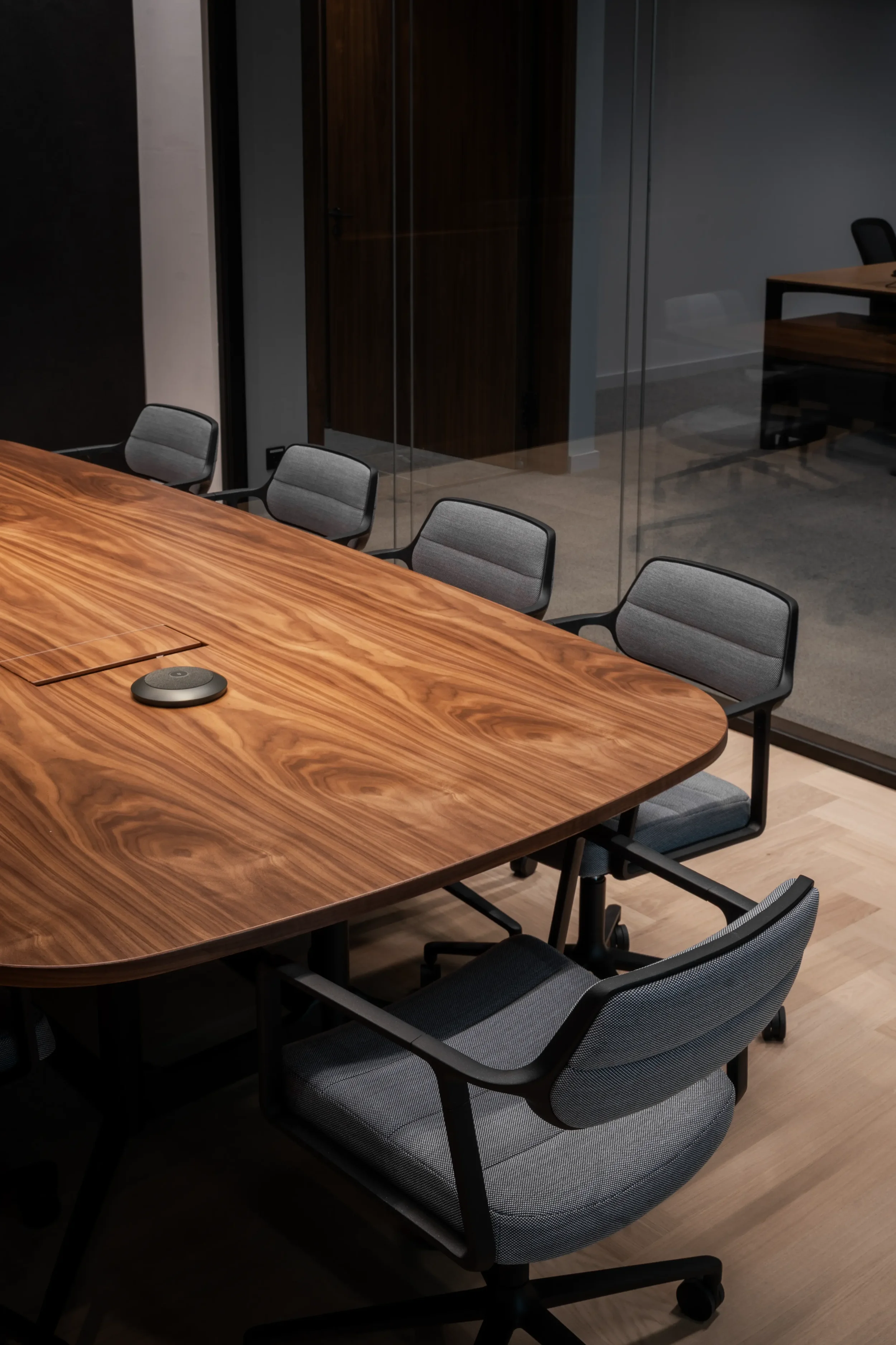 Ein moderner Konferenztisch aus Holz mit mehreren schwarzen und grauen Bürostühlen, eine Konferenztechnik auf dem Tisch, im Hintergrund eine gläserne Trennwand und eine robuste Holz-Schrankwand im Büro.