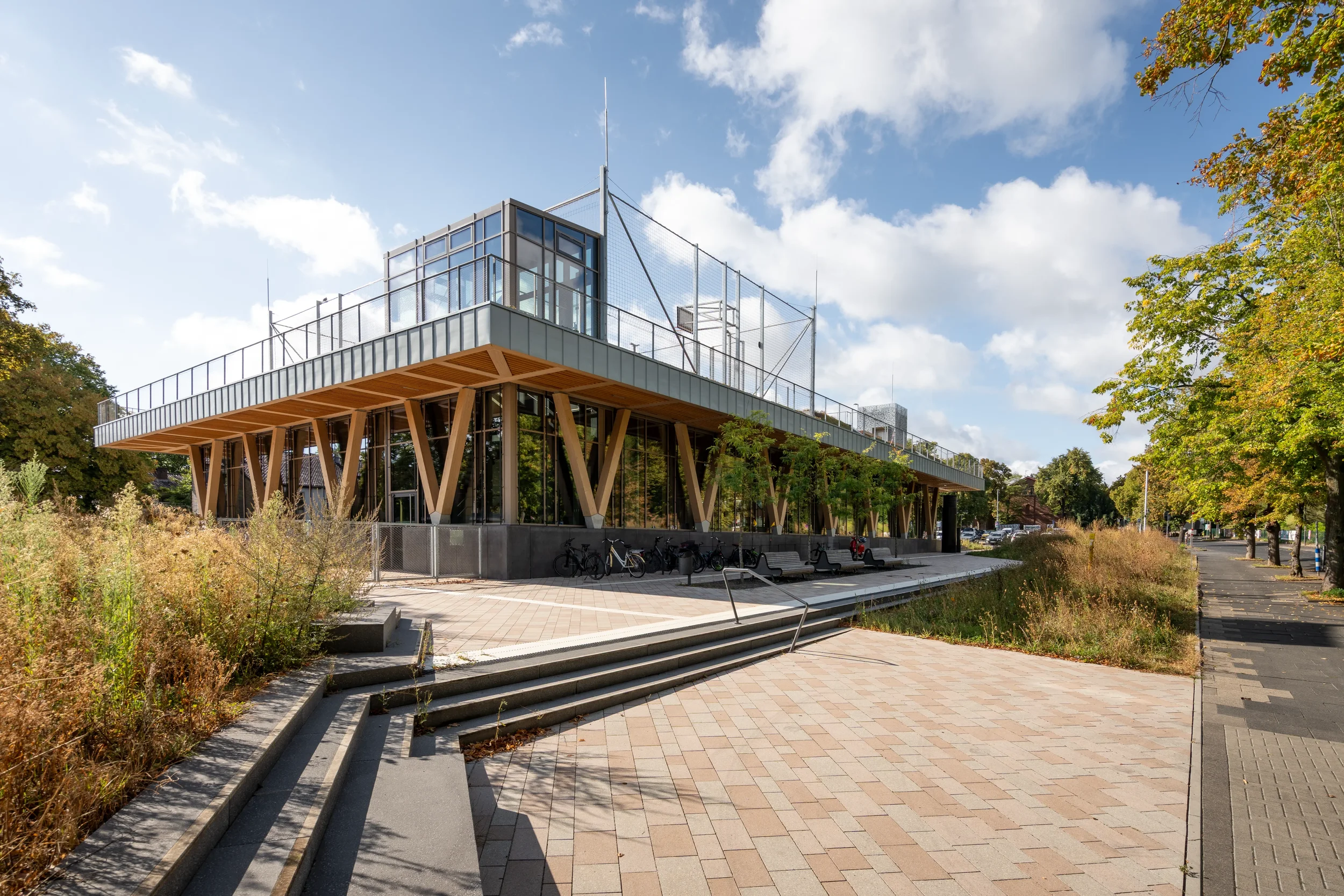 Modernes zweistöckiges Gebäude mit Holzbalken, große Fenster und Außenterrasse, umgeben von Bäumen und Garten, bei sonnigem Himmel.