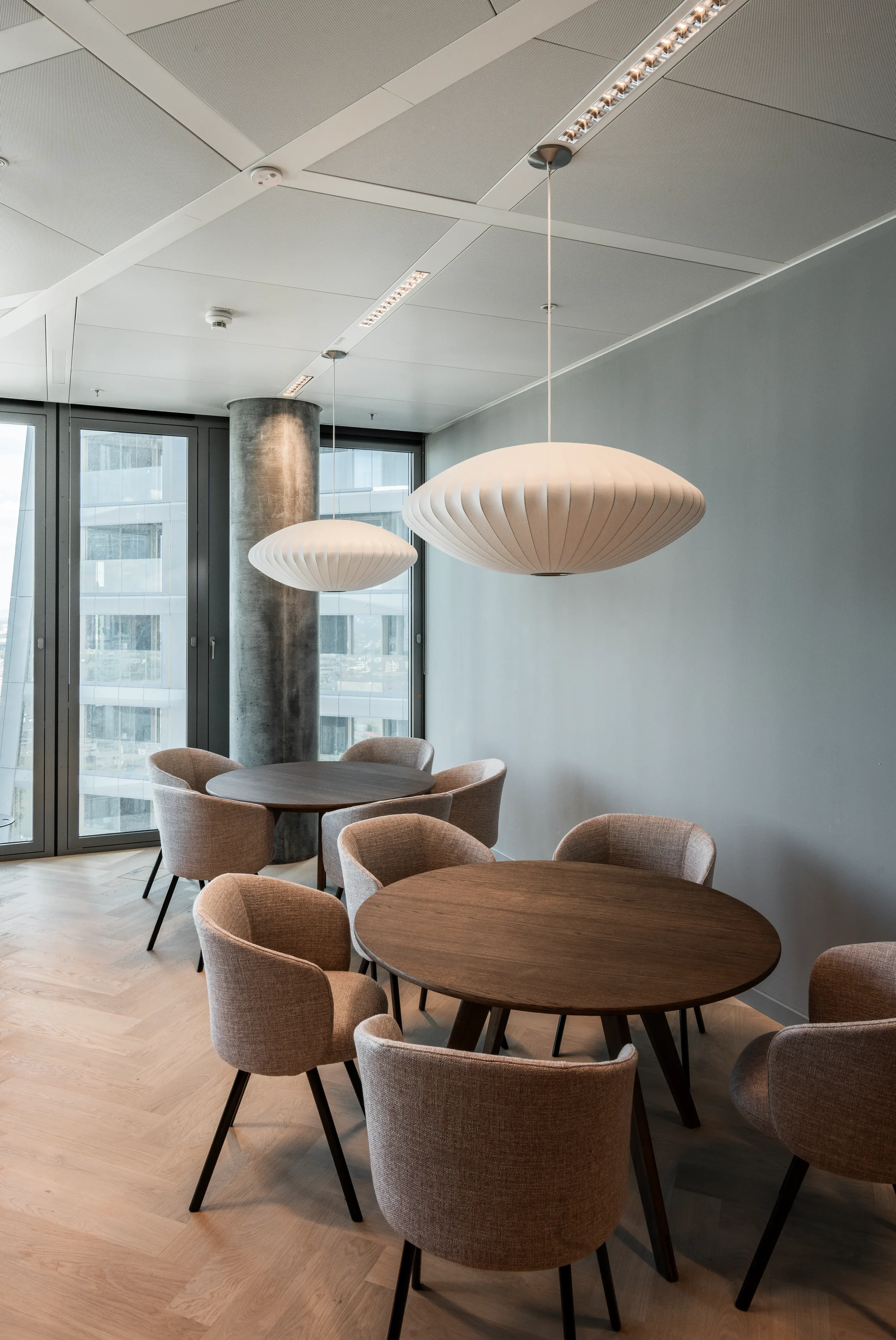 Innenraum mit runden Holztischen, gepolsterten Stühlen und two großen, hängenden Lampen, große Fenster mit Blick auf städtische Gebäude.