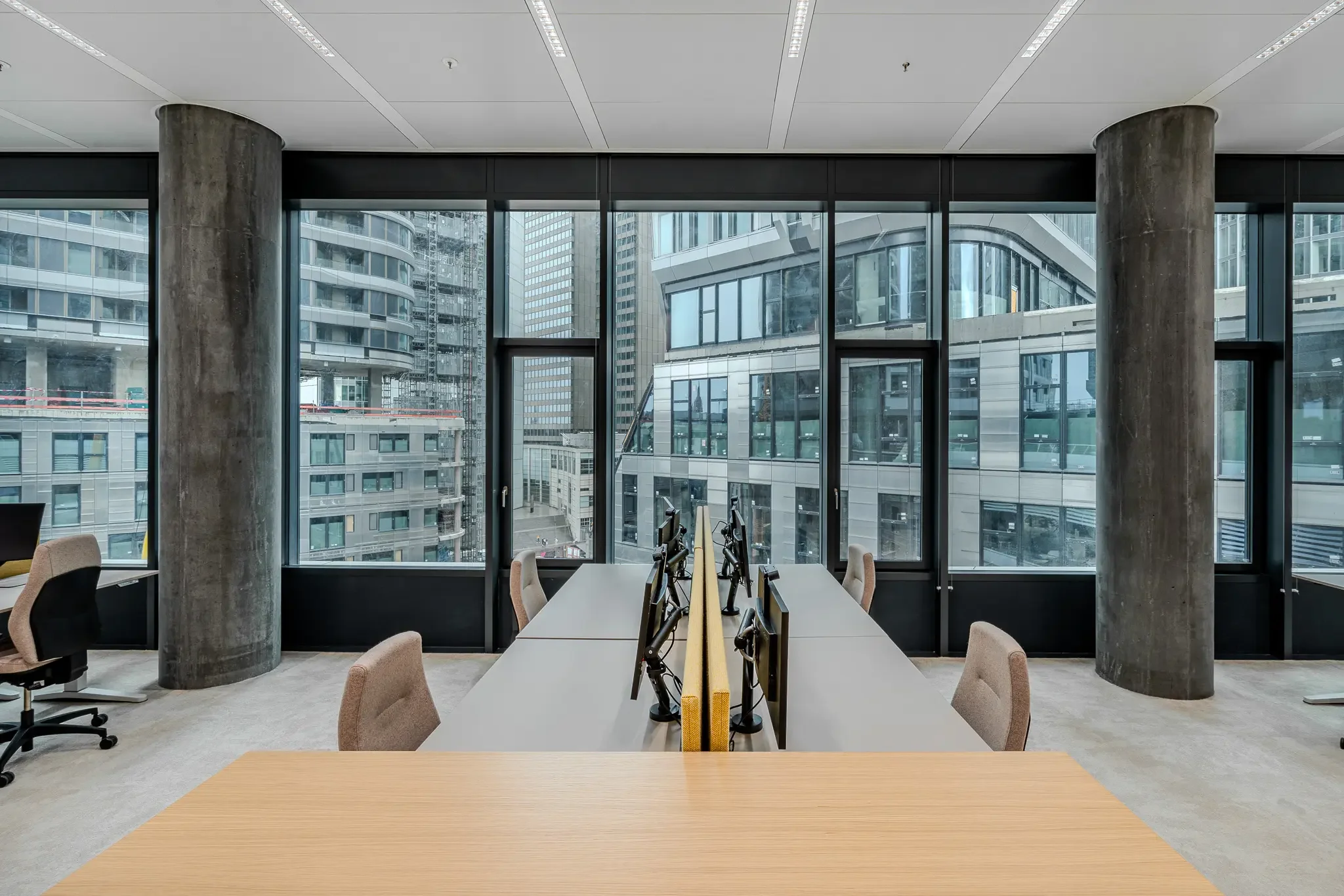 Ein moderner Büroarbeitsraum mit großen Fenstern, Cityblick, Schreibtisch mit Computern, Bürostühlen und grauen Sitzgelegenheiten.
