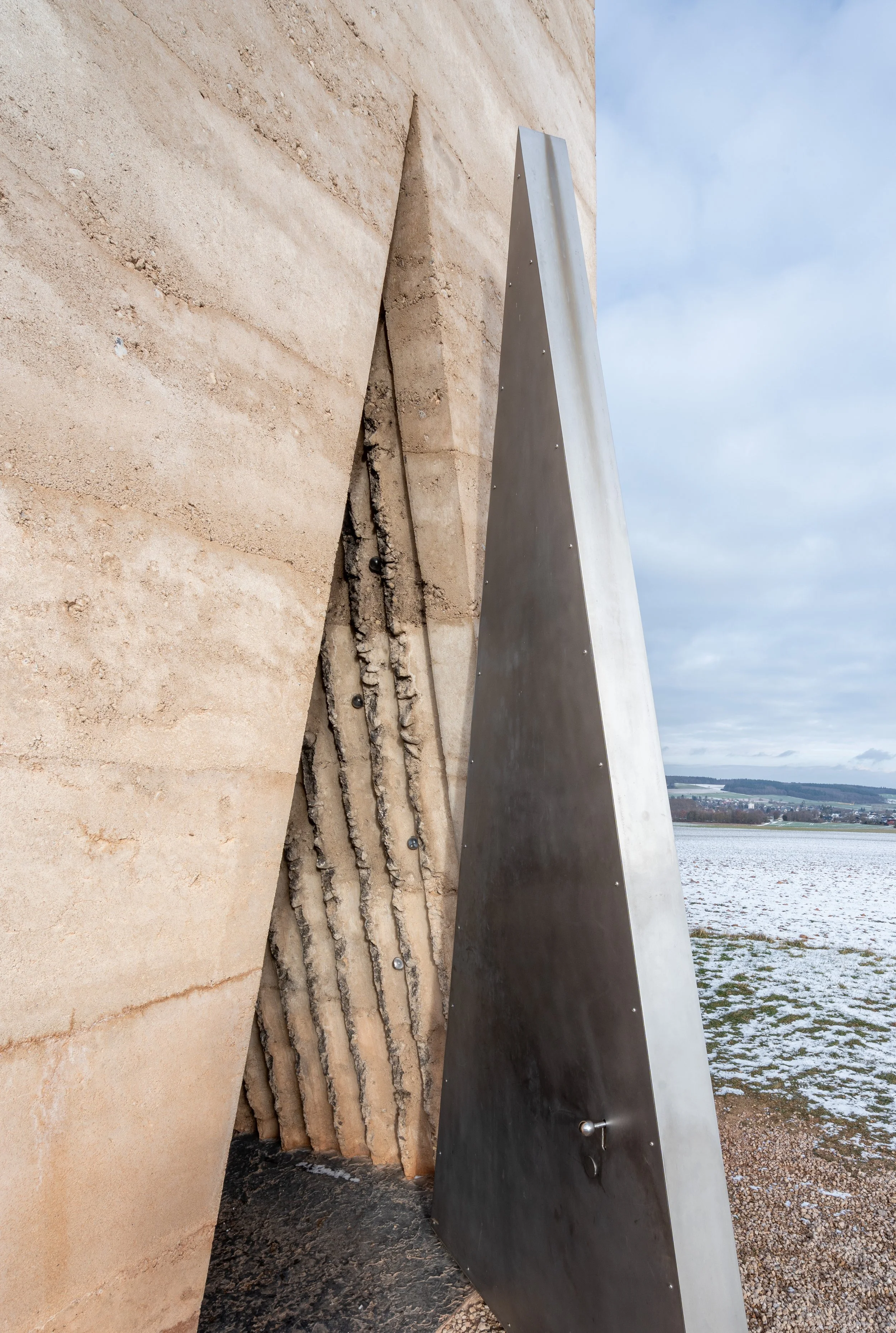 Moderne Skulptur aus Stahl und Stein vor einer winterlichen Landschaft mit Wasser und Feld im Hintergrund.