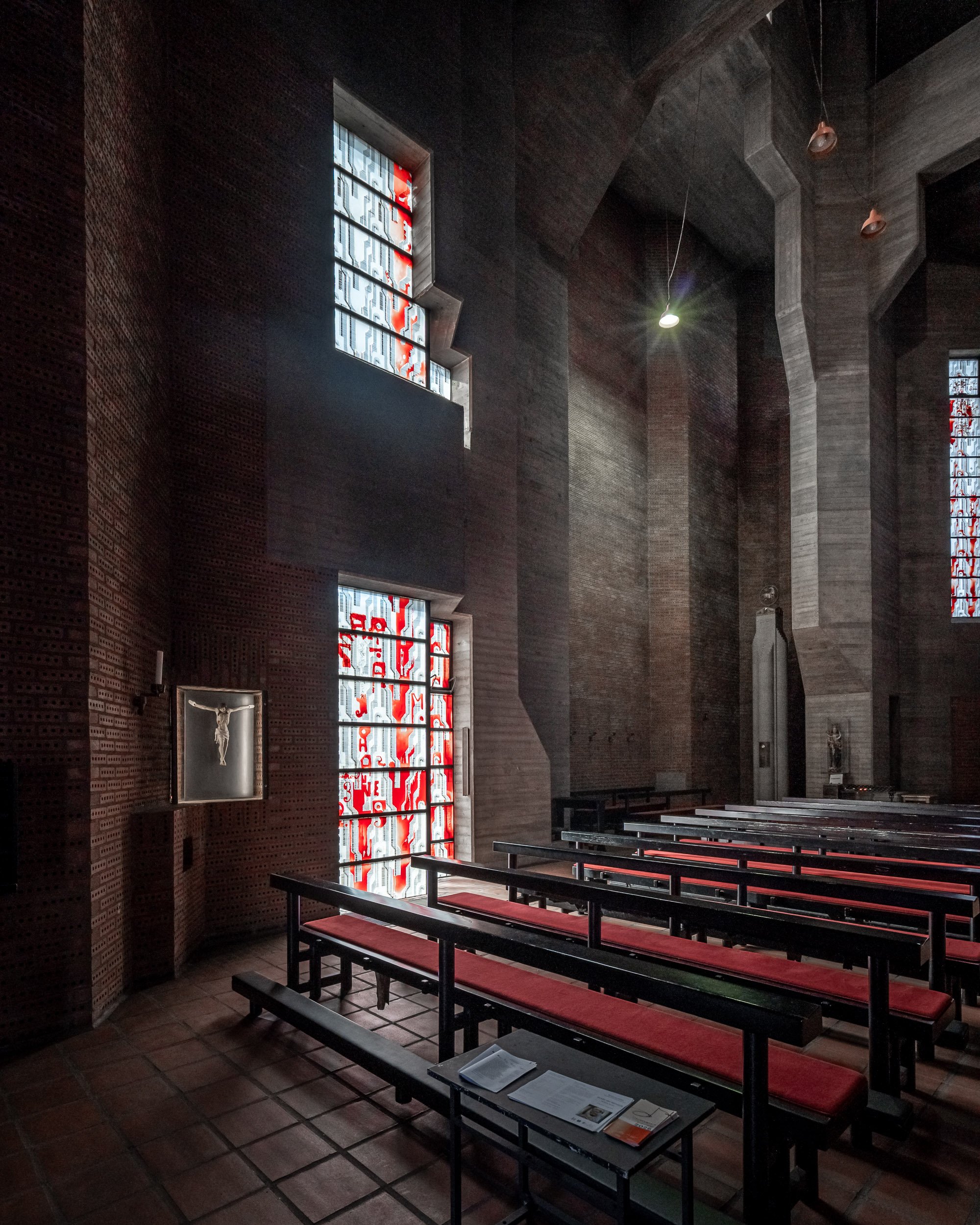 Innenraum einer Kirche mit hohen Wänden aus rotem Ziegelstein, Kirchenbänken mit roten Sitzpolstern, großen bunten Kirchenfenstern und einigen Skulpturen.