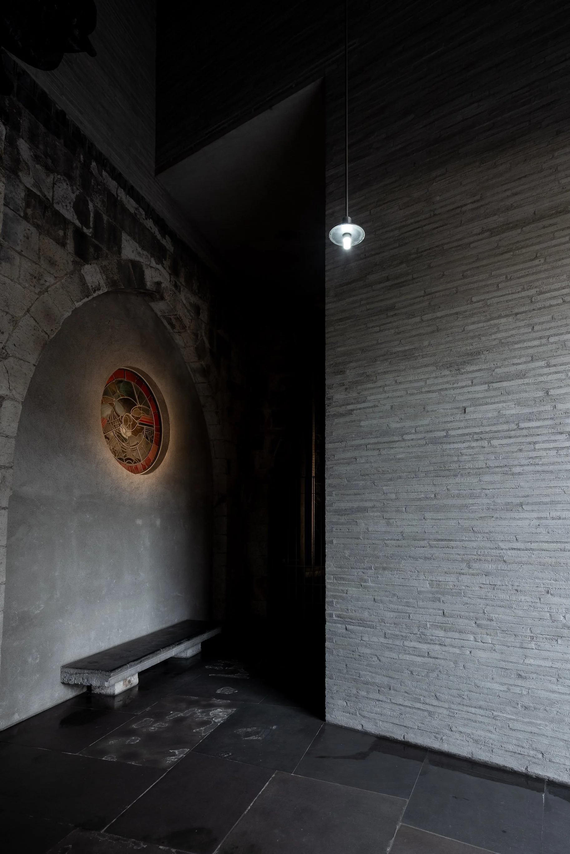 Innenaufnahme eines dunklen Raums mit einer runden Uhr an einer Wand, einem Steinbank und modernen Wand- und Deckenflächen.