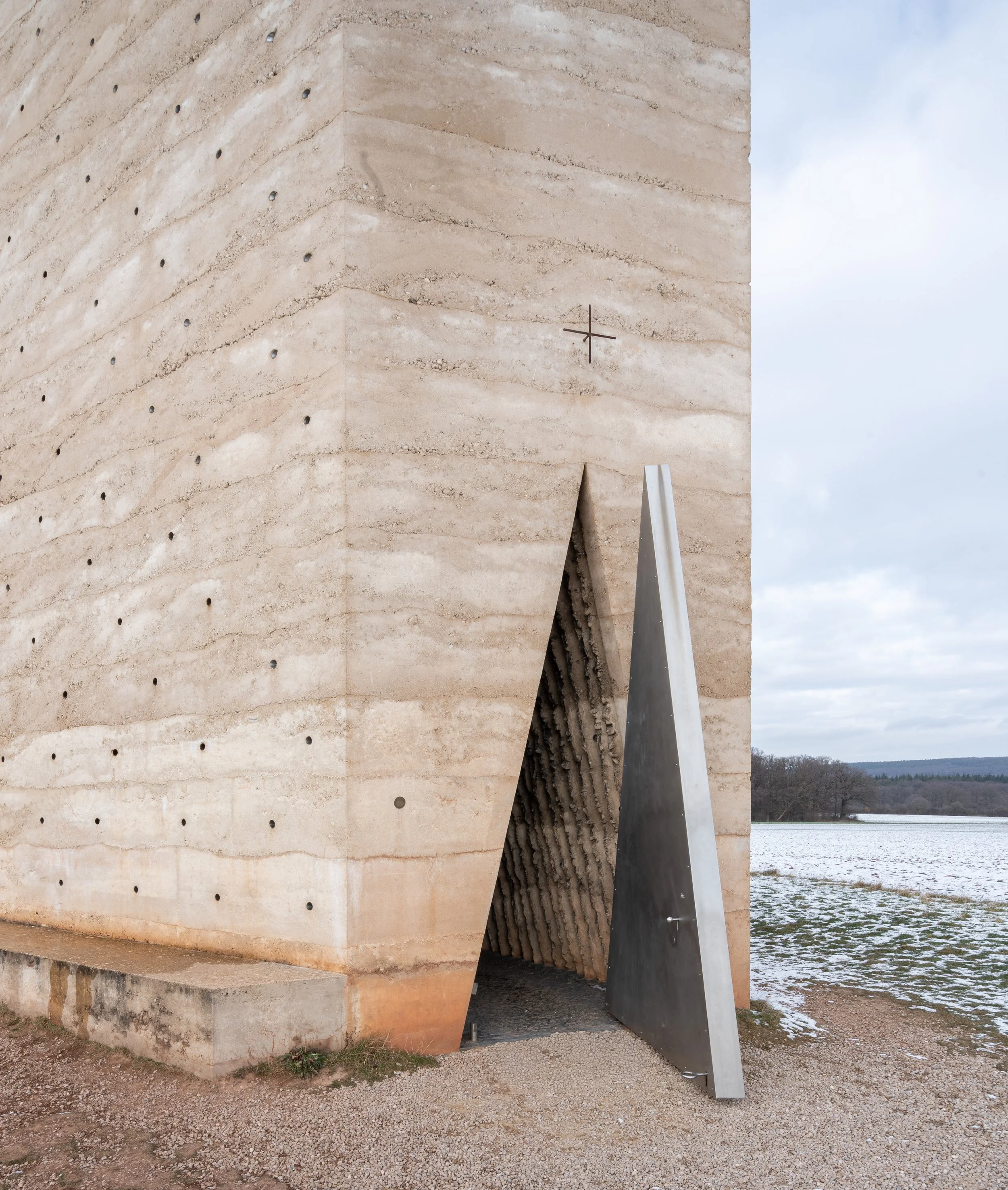Moderne Kirche aus Sandstein mit einem offenen metallischen Eingang und einer kleinen Eisenkreuz auf der Außenwand, auf einer ländlichen, schneebedeckten Landschaft bei bewölktem Himmel.