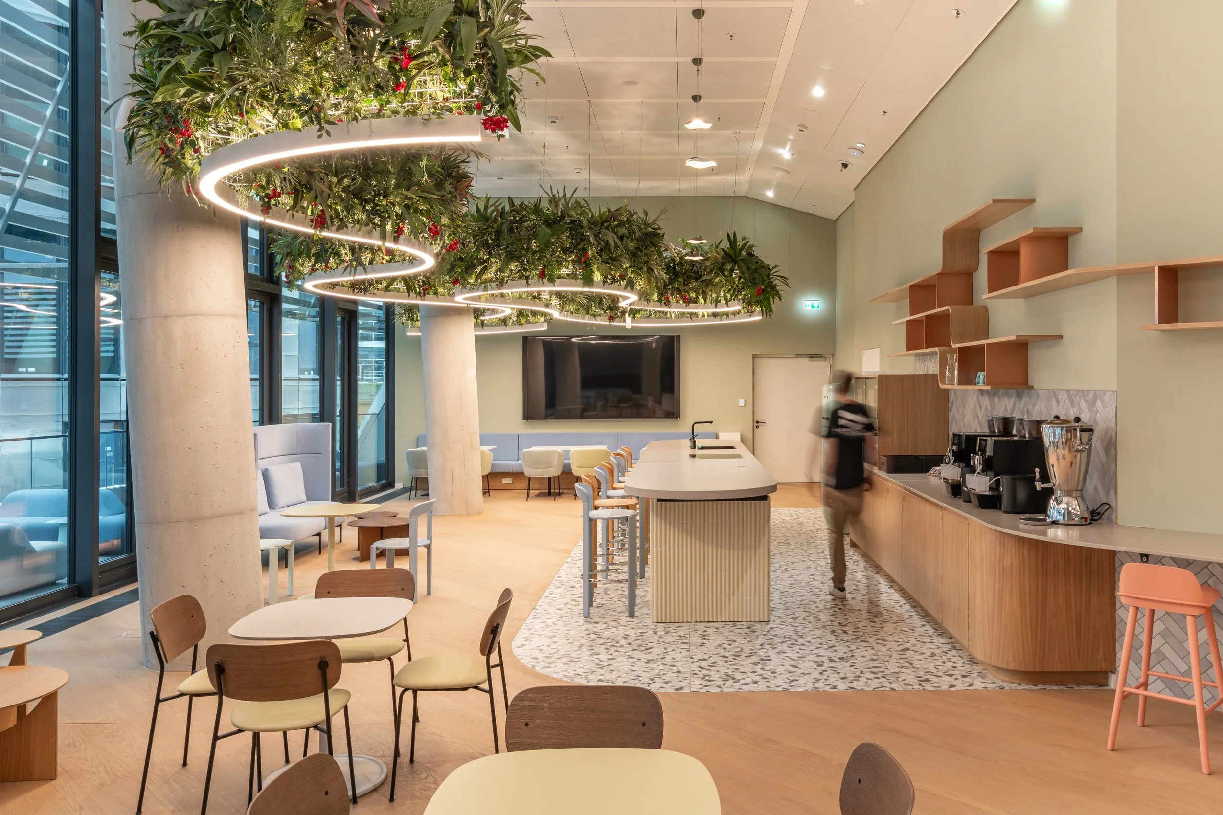 Moderne Cafeteria mit Barista-Bereich, großen Fenstern, Pflanzenhängetröge mit Lichteranzug, gemütlichen Sitzgelegenheiten, einem großen Bildschirm, Holzwänden und einem Kaffeeautomaten.