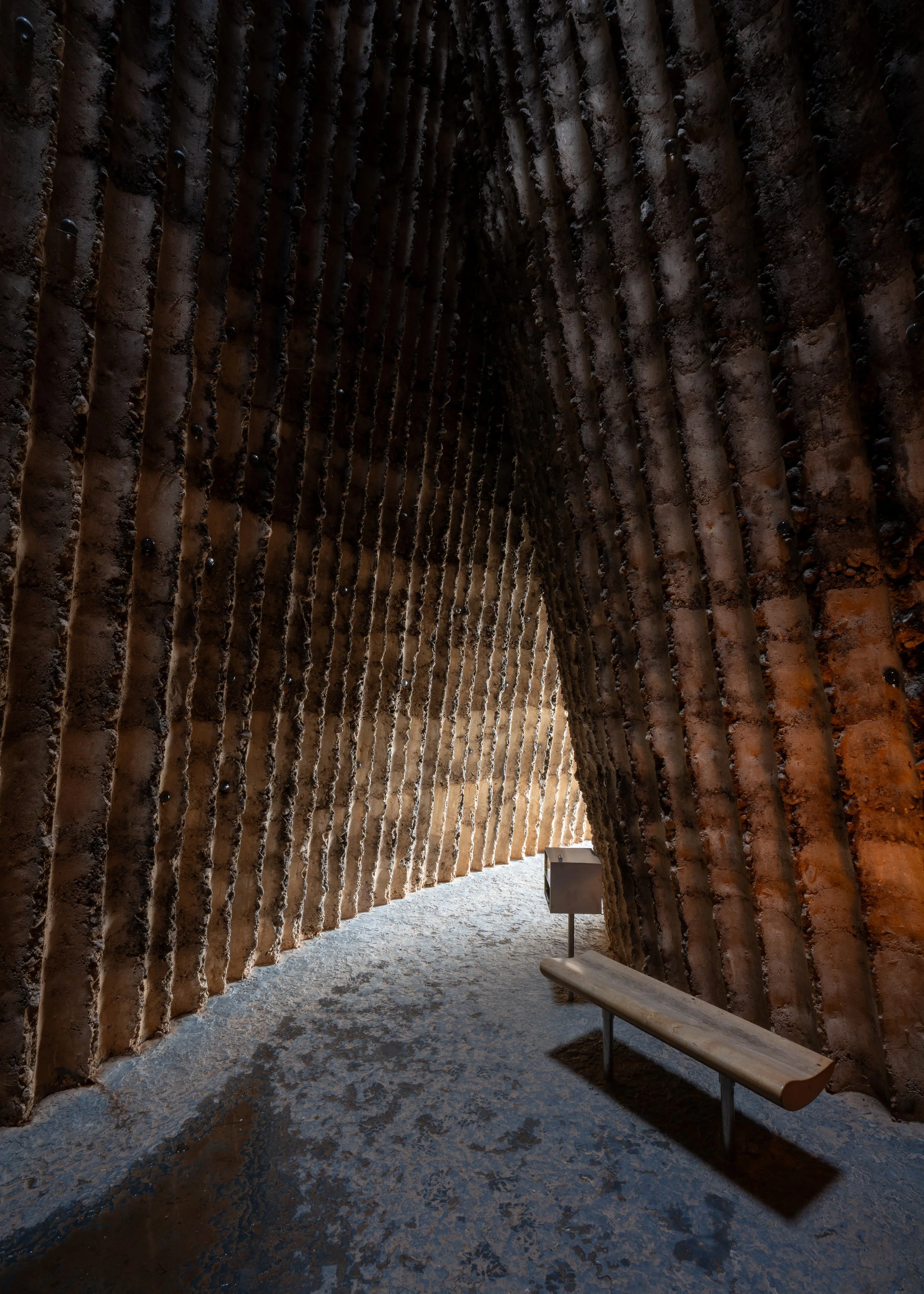 Innenraum eines Kunstwerks, mit großen, gebogenen, strukturierten Wänden aus Salz oder Kalkstein, ein Holzbank und ein kleines Informationsschild davor.
