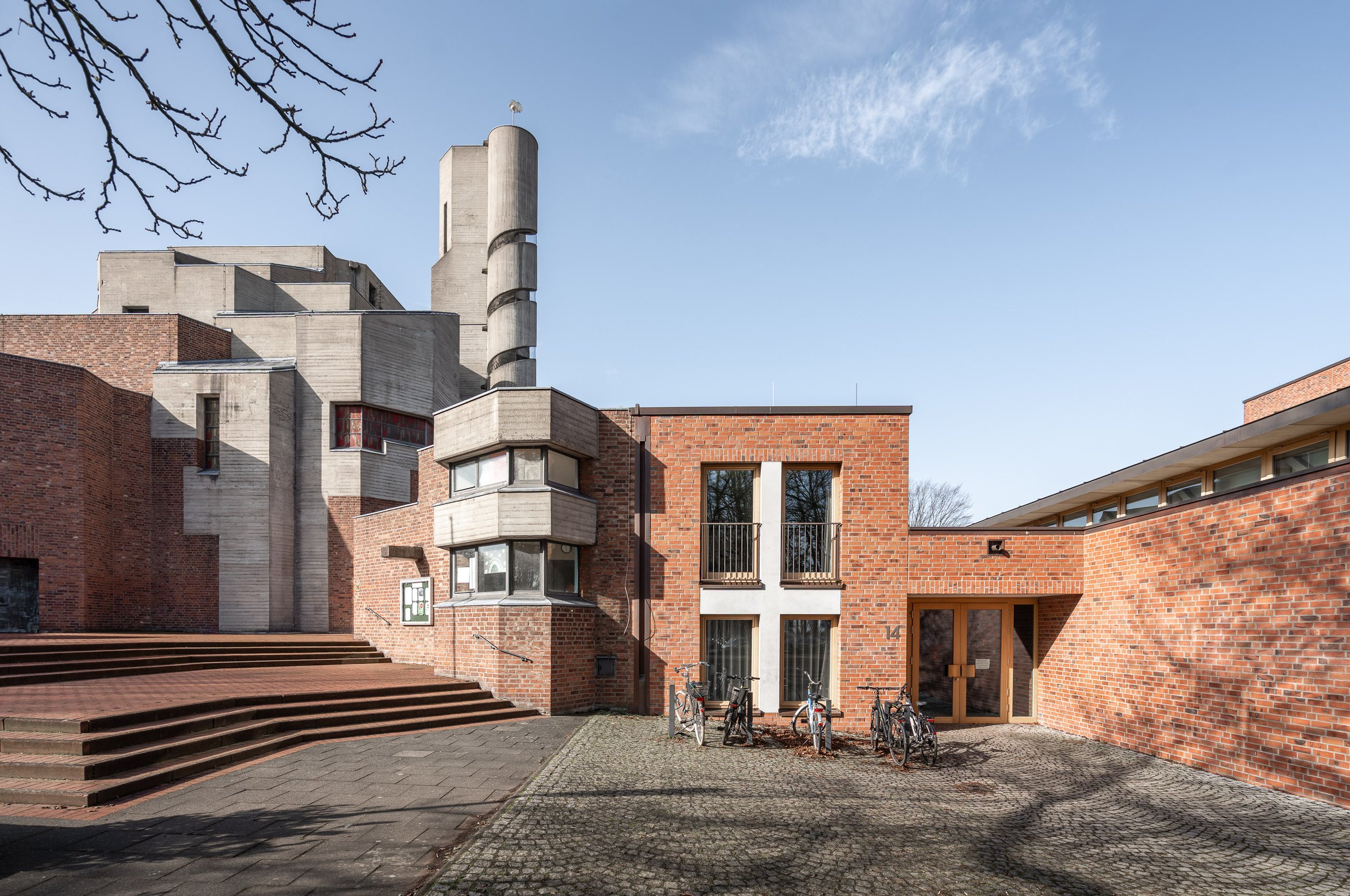 Modernes Gebäude mit roten Ziegeln, Treppen, Fahrrädern vor dem Eingang, blauer Himmel im Hintergrund.