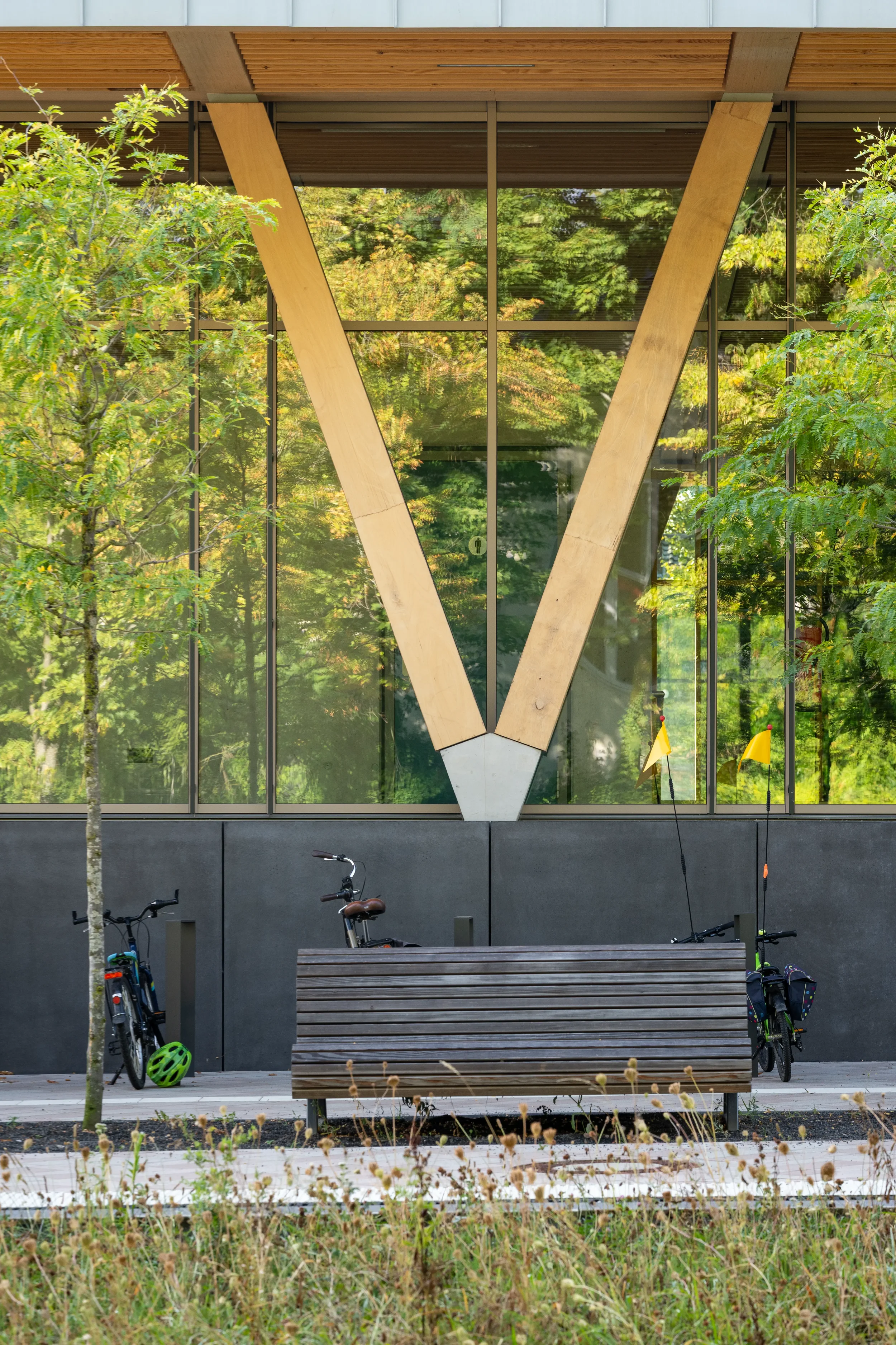 Detail Architekturfotografie Kultur- und Sporthalle in Alfter, Moderne Gebäudefassade mit großen Glasfenstern, Holzträgern in V-Form, Bänken, Fahrrädern, Helmen und kleinen Flaggen vor einem grünen, bewaldeten Hintergrund.